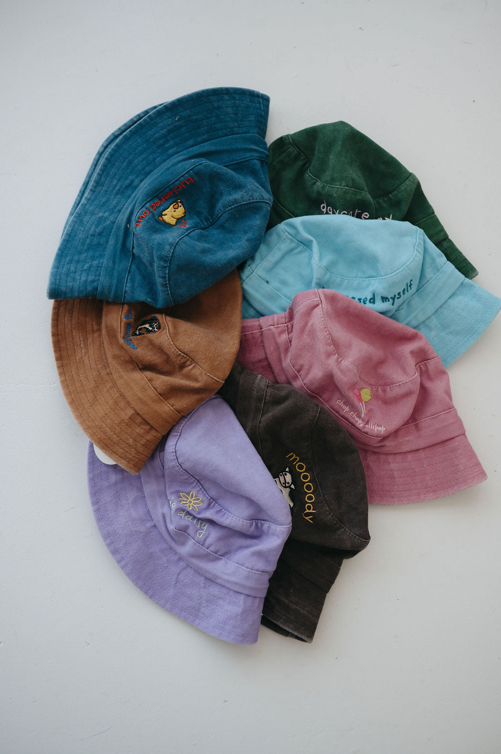A group of seven toddler daycare washed cotton bucket hats in various colours (light blue, green, brown, dark blue, pink, purple, dark brown) arranged in a circle on a plain white background.