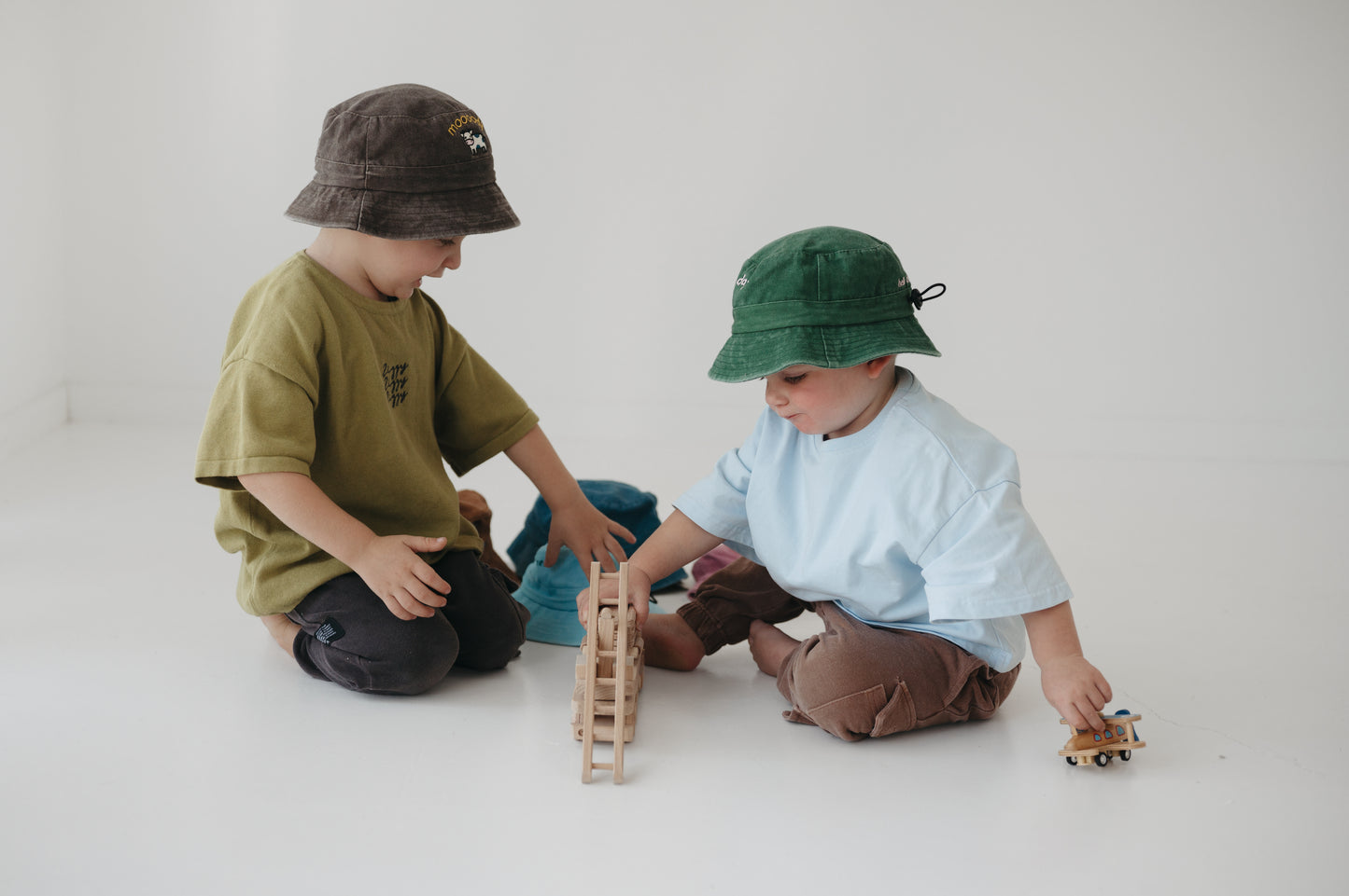 Two toddlers sitting on a white floor playing with wooden blocks and a toy car, wearing a dark brown washed cotton daycare bucket hat and a green washed cotton daycare bucket hat with casual t-shirts in a bright, minimalist room.