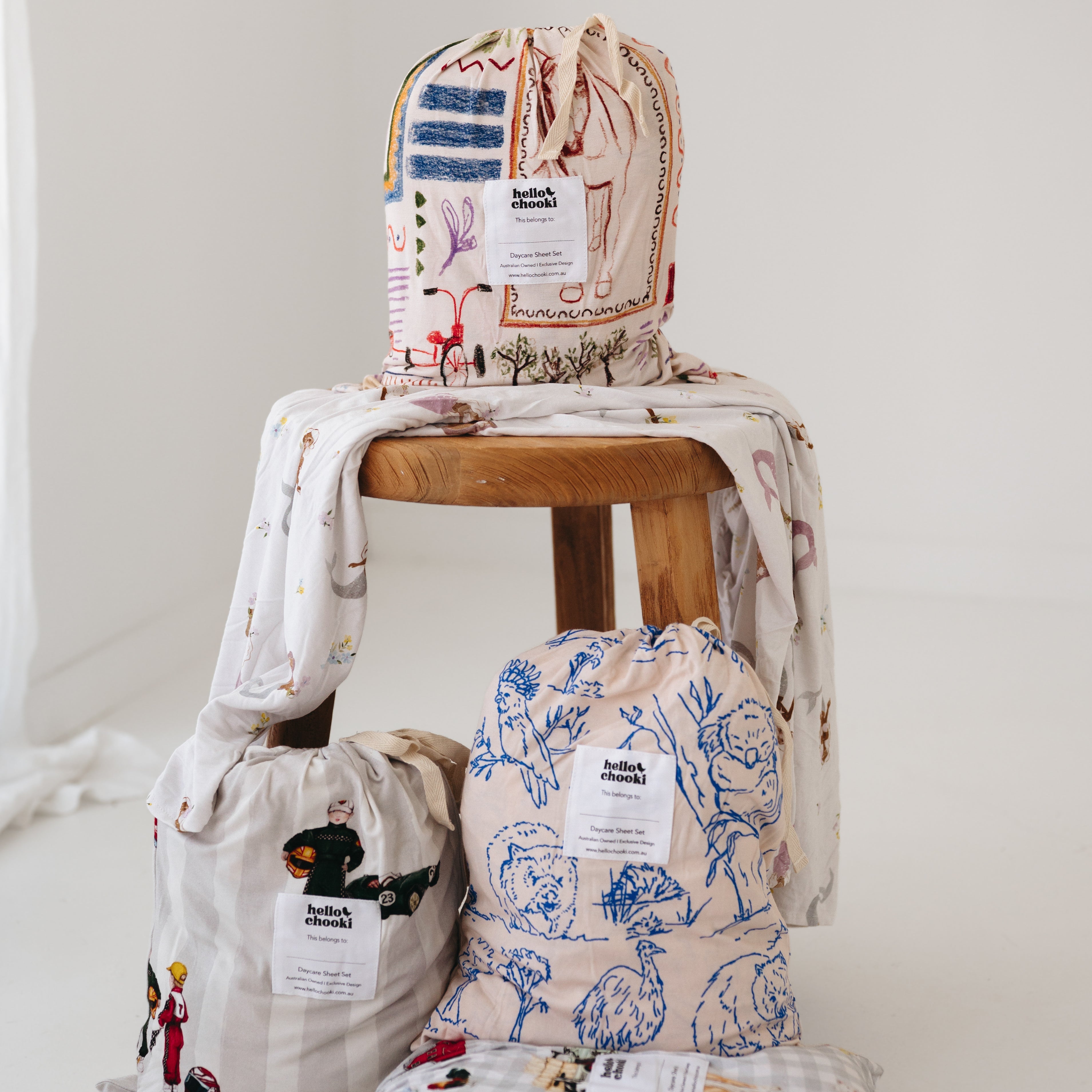 Beige and multicolour daycare sheet set with hand drawn geometric shapes featuring a horse, vintage bike, tent, campfire, fishing, native trees and animals, packaged in its drawstring bag on a wooden stool, white background.