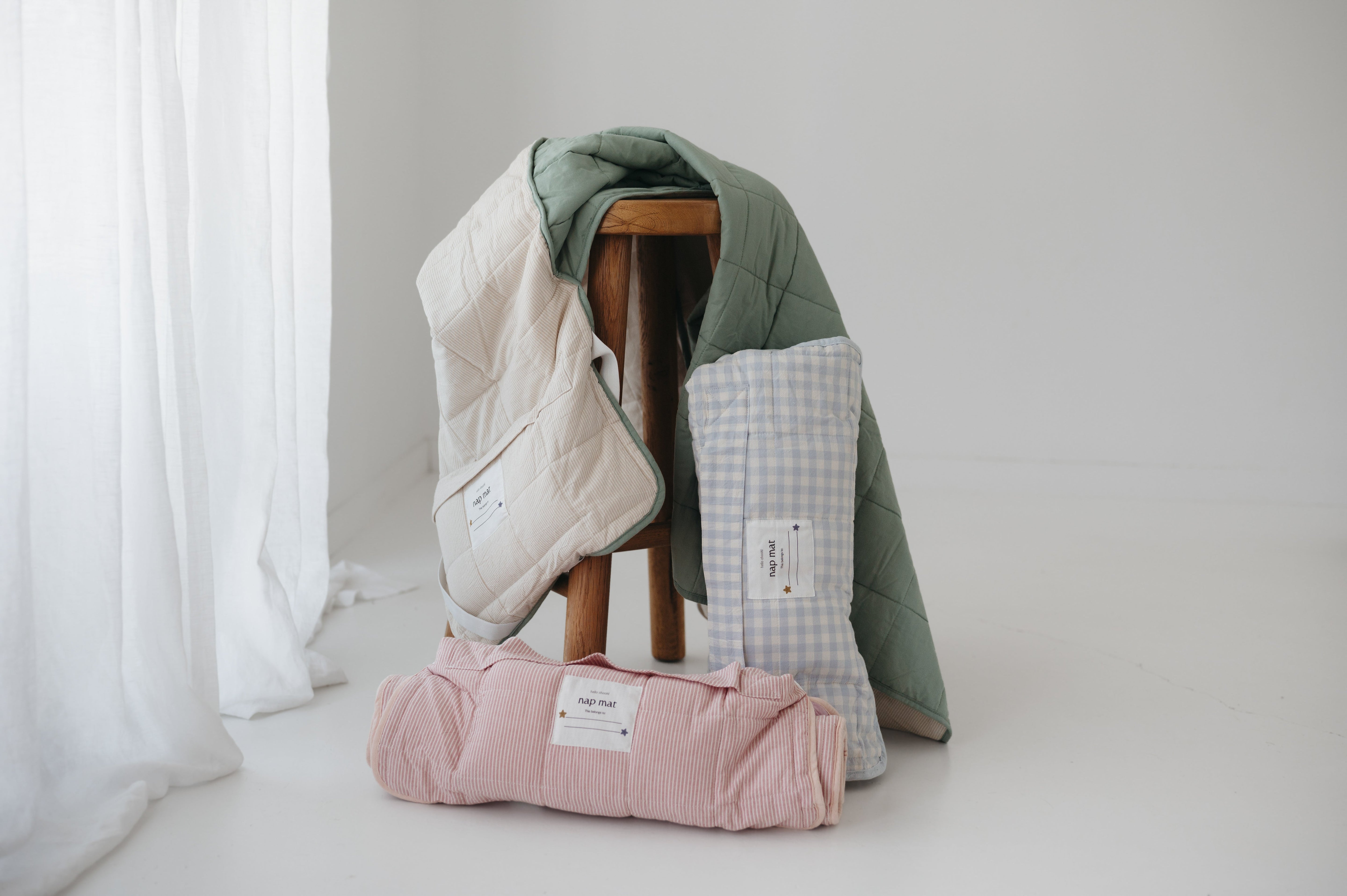 Olive green and beige stripe quilted daycare nap mat with attached dark green pillowcase, pillow insert, name tag, and carry handle, draped over a timber stool with alternative colours from the collection.