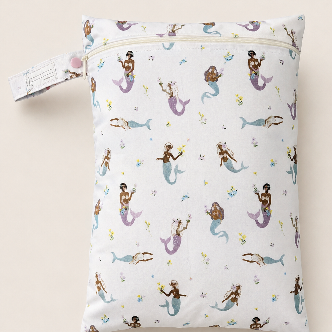 Rectangle zippered daycare wet bag with a repeating purple and blue mermaid pattern on white, shown against a light beige backdrop. Features a pastel purple button for attaching to a bag or pram, plus a name tag to prevent mixups.