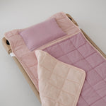 A soft purple and dusty pink stripe quilted daycare bedding nap mat with a soft peach underside on quilt. It has a built in matching pillowcase, pillow insert, name tag and carry handle. The daycare bedding nap mat is laid out on a daycare stacker bed frame against a white background.