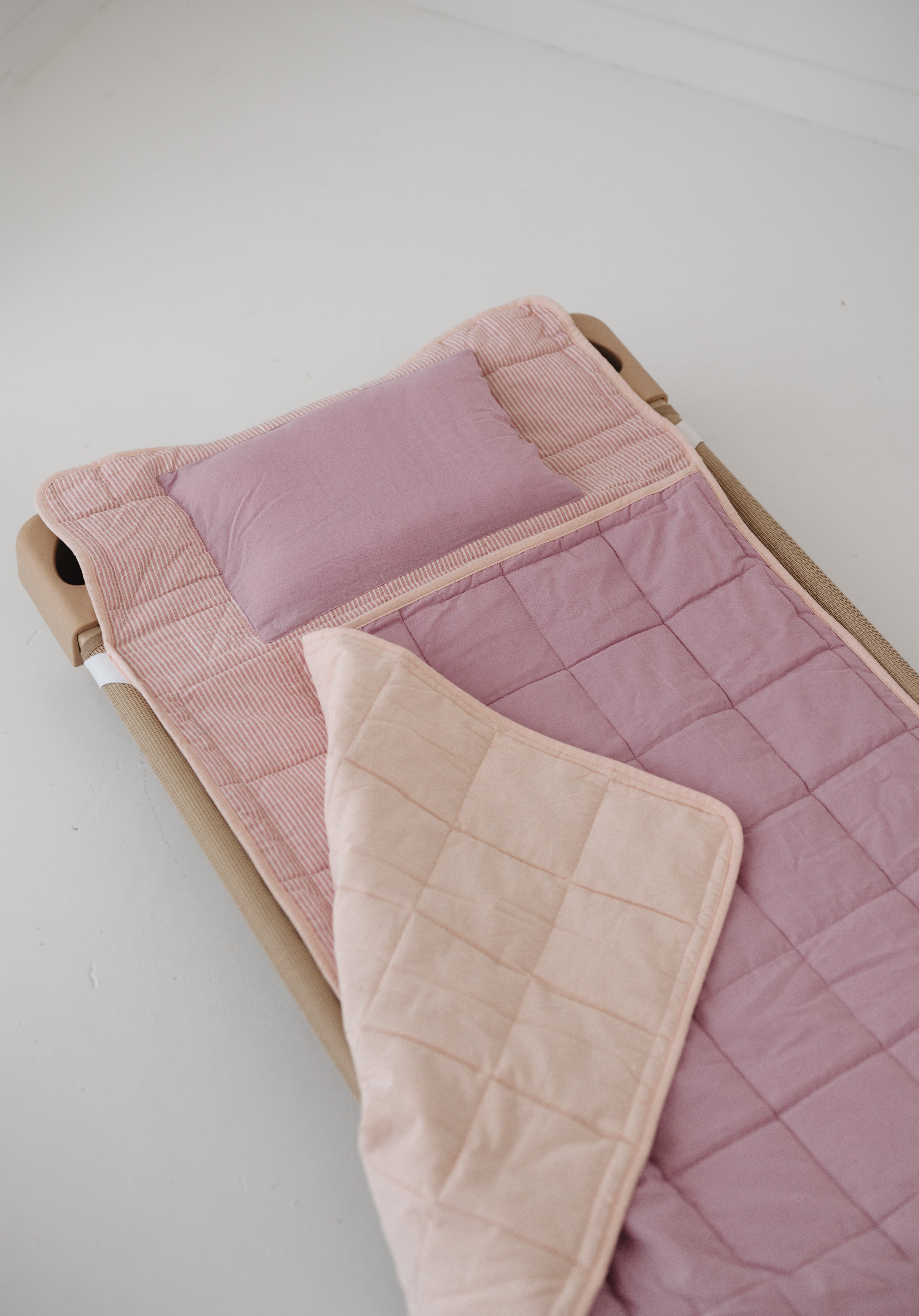 A soft purple and dusty pink stripe quilted daycare bedding nap mat with a soft peach underside on quilt. It has a built in matching pillowcase, pillow insert, name tag and carry handle. The daycare bedding nap mat is laid out on a daycare stacker bed frame against a white background.