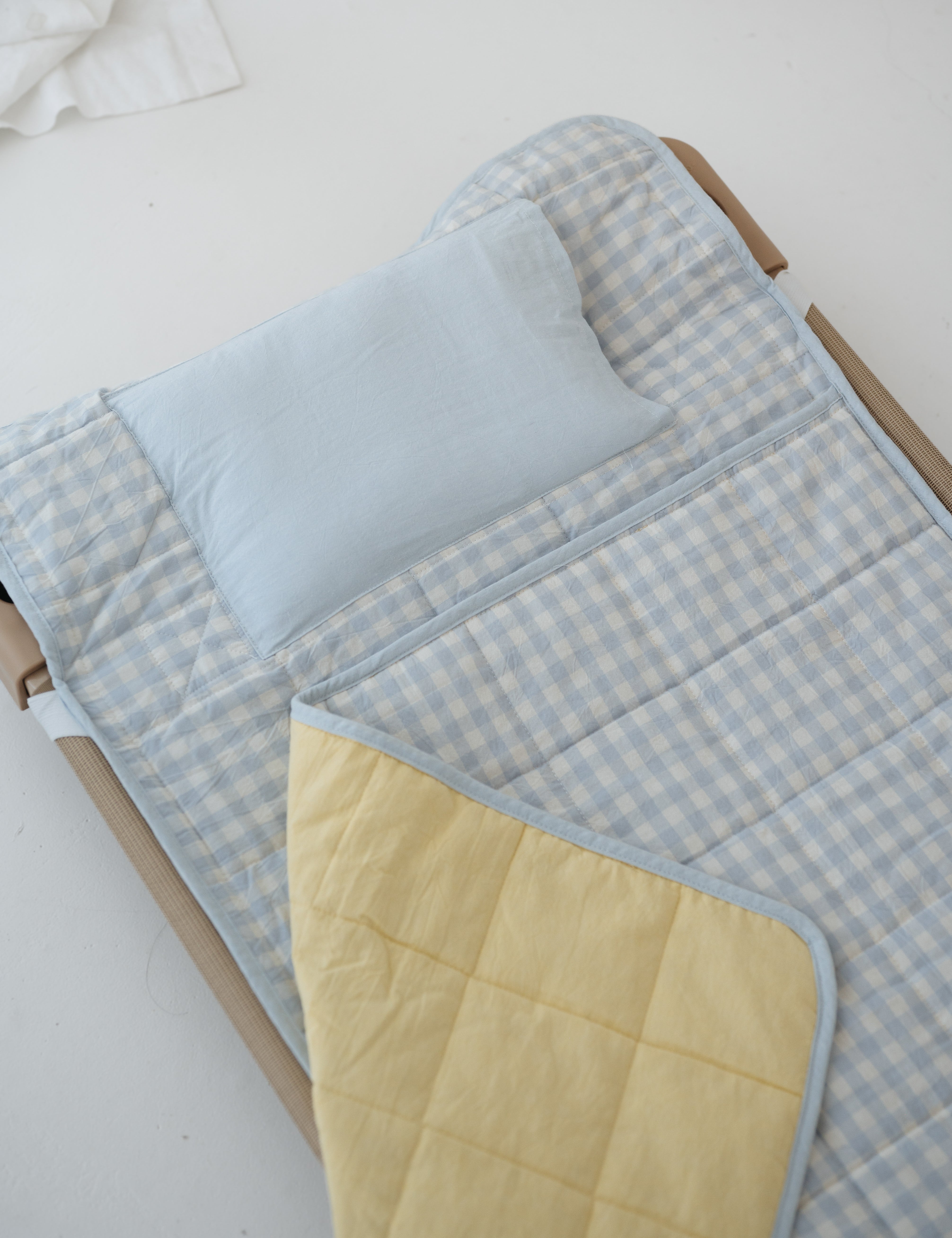 A light blue and white gingham quilted daycare bedding nap mat with soft yellow on the underside of the blanket. It has a built in matching pillowcase, pillow insert, name tag and carry handle. The daycare bedding nap mat is laid out on a daycare stacker bed frame against a white background.