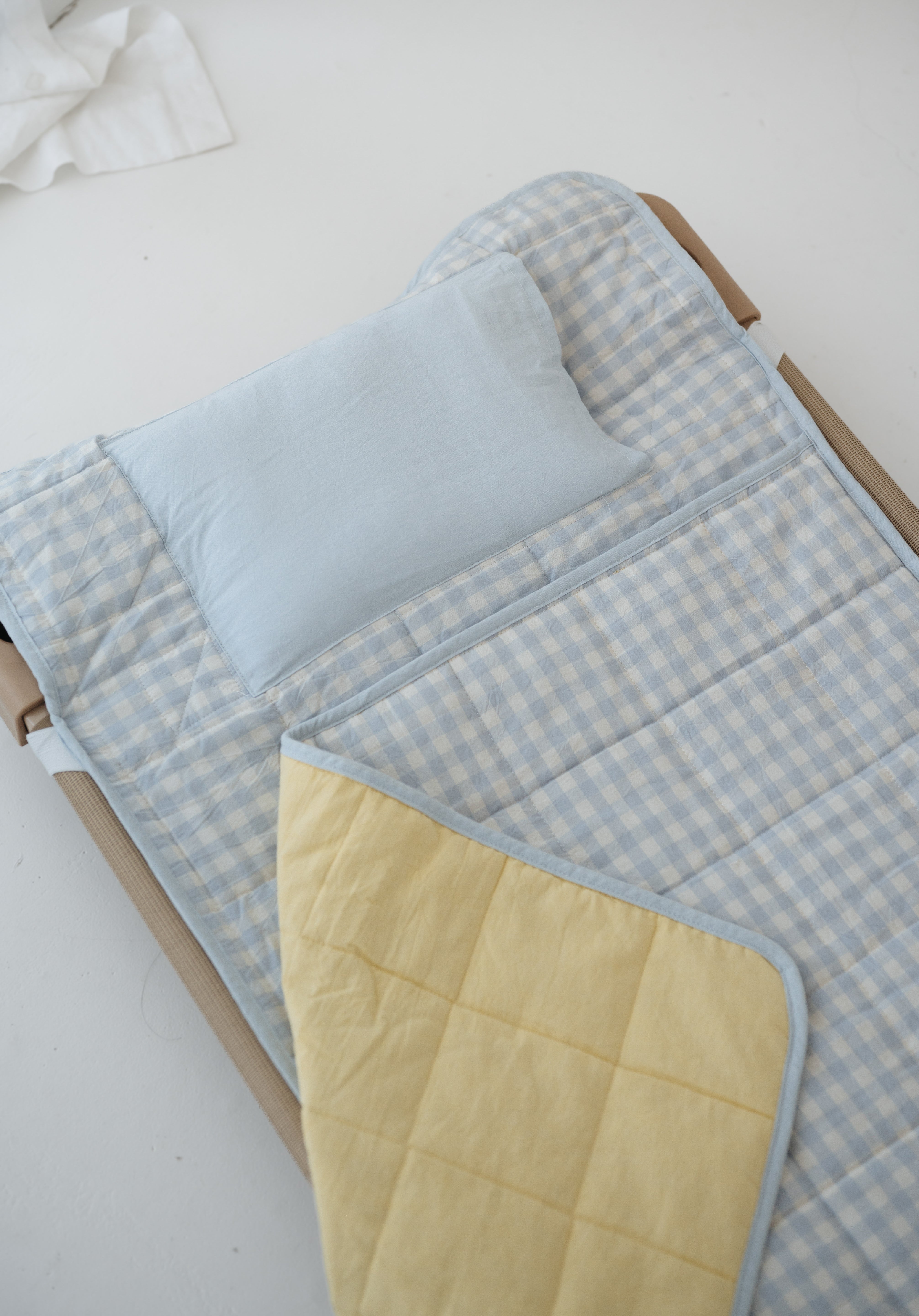 A light blue and white gingham quilted daycare bedding nap mat with soft yellow on the underside of the blanket. It has a built in matching pillowcase, pillow insert, name tag and carry handle. The daycare bedding nap mat is laid out on a daycare stacker bed frame against a white background.