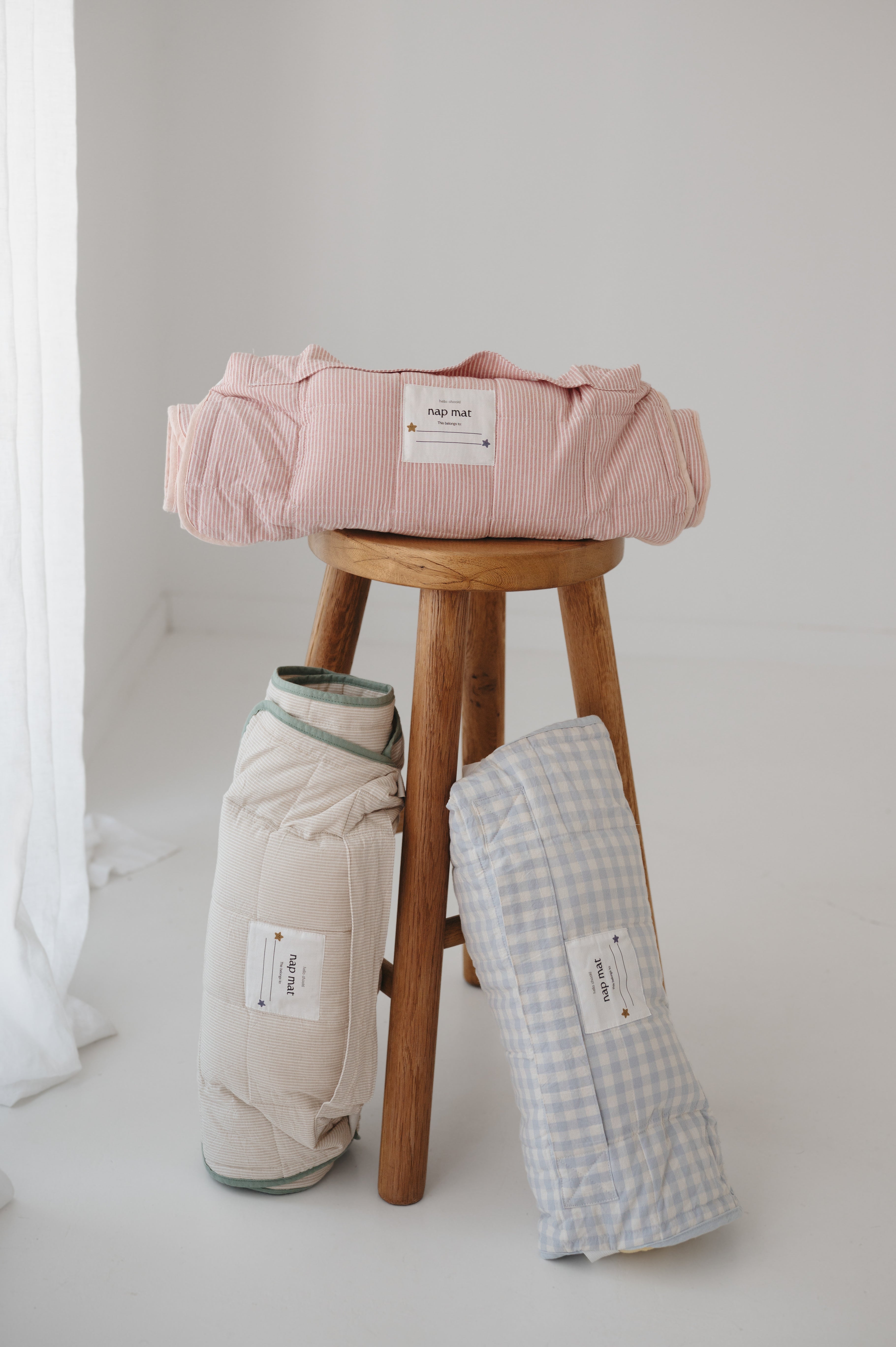 A soft purple and dusty pink stripe quilted daycare bedding nap mat with a soft peach underside on quilt. It has a built in matching pillowcase, pillow insert, name tag and carry handle. The daycare bedding nap mat is rolled up onto of a wooden stool next to alternative colours within the daycare bedding nap mat collection, photographed with a white background.