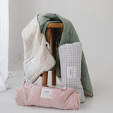 A light blue and white gingham quilted daycare bedding nap mat with soft yellow on the underside of the blanket. It has a built in matching pillowcase, pillow insert, name tag and carry handle. The daycare bedding nap mat is rolled up against a wooden stool with alternative daycare bedding nap mats from the Hello Chooki collection, photographed with a white background.