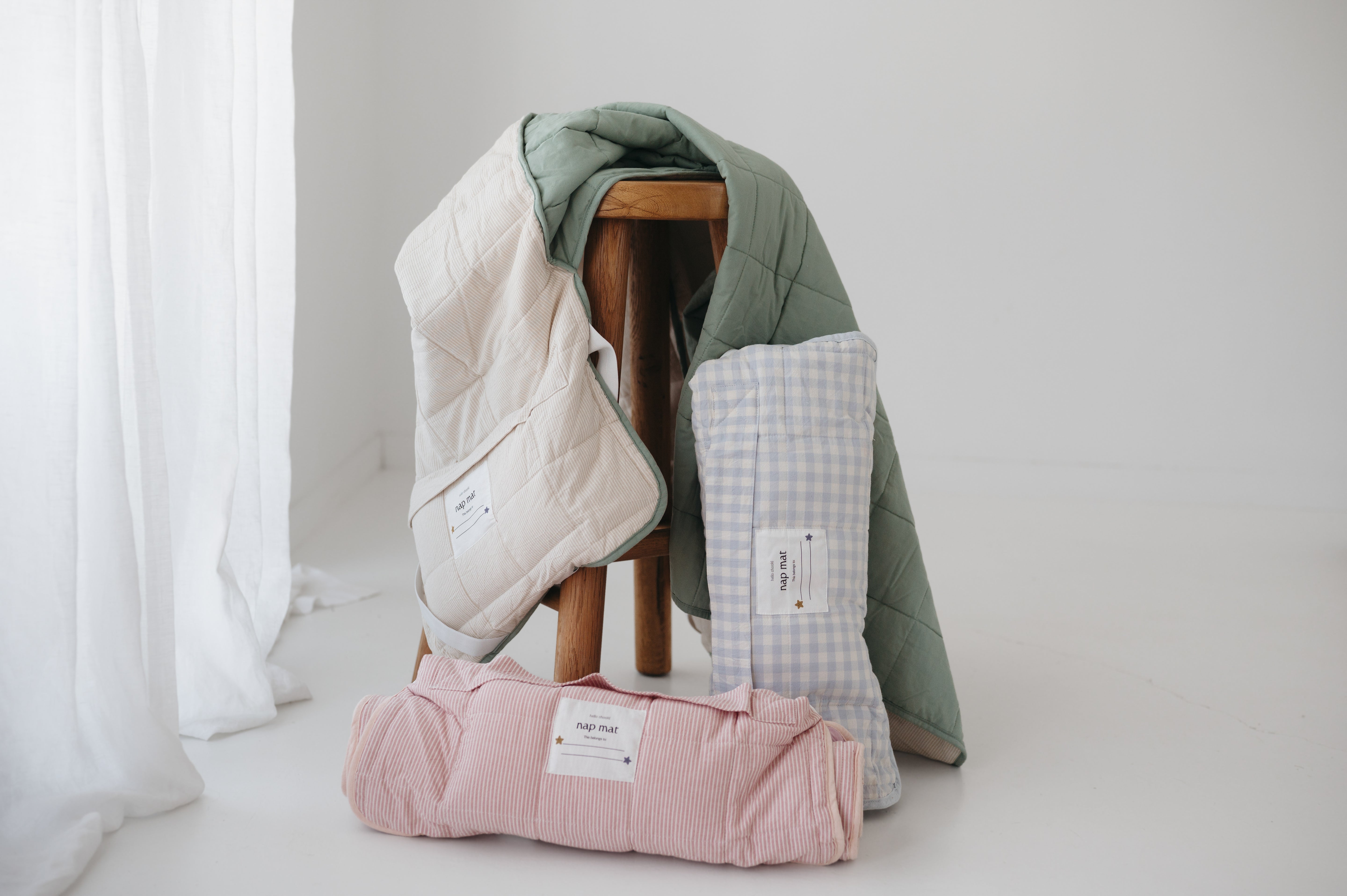 A light blue and white gingham quilted daycare bedding nap mat with soft yellow on the underside of the blanket. It has a built in matching pillowcase, pillow insert, name tag and carry handle. The daycare bedding nap mat is rolled up against a wooden stool with alternative daycare bedding nap mats from the Hello Chooki collection, photographed with a white background.