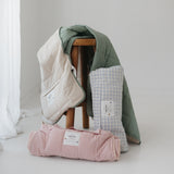 A olive green and beige stripe quilted daycare bedding nap mat with a dark green pillowcase attached. It has a built in matching pillowcase, pillow insert, name tag and carry handle. The daycare bedding nap mat is draped over a timber stool with alternative colours next to it from the daycare bedding nap mat collection.
