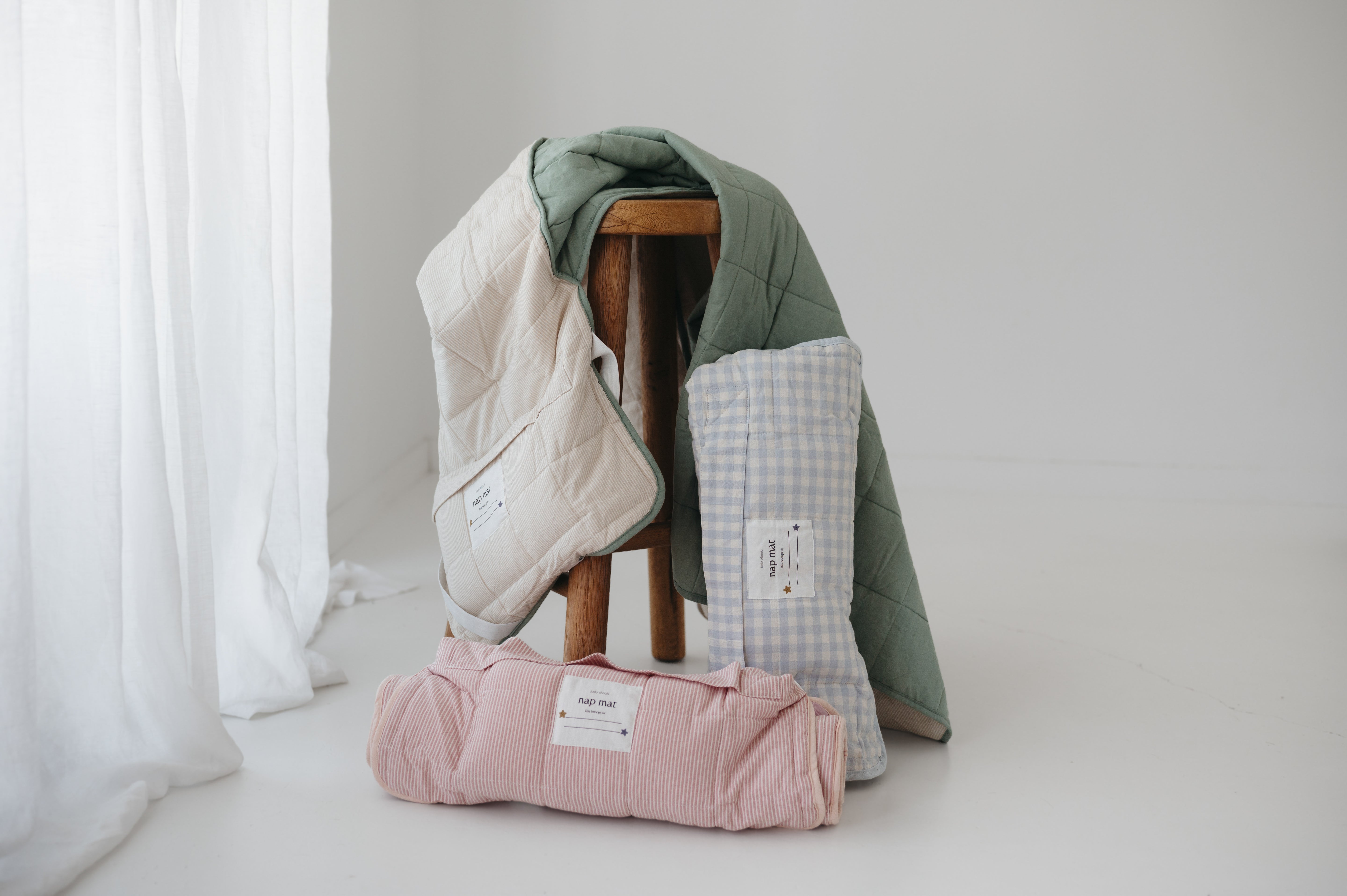 A olive green and beige stripe quilted daycare bedding nap mat with a dark green pillowcase attached. It has a built in matching pillowcase, pillow insert, name tag and carry handle. The daycare bedding nap mat is draped over a timber stool with alternative colours next to it from the daycare bedding nap mat collection.