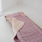 A soft purple and dusty pink stripe quilted daycare bedding nap mat with a soft peach underside on quilt. It has a built in matching pillowcase, pillow insert, name tag and carry handle. The daycare bedding nap mat is laid out on a daycare stacker bed frame against a white background.