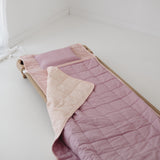 A soft purple and dusty pink stripe quilted daycare bedding nap mat with a soft peach underside on quilt. It has a built in matching pillowcase, pillow insert, name tag and carry handle. The daycare bedding nap mat is laid out on a daycare stacker bed frame against a white background.