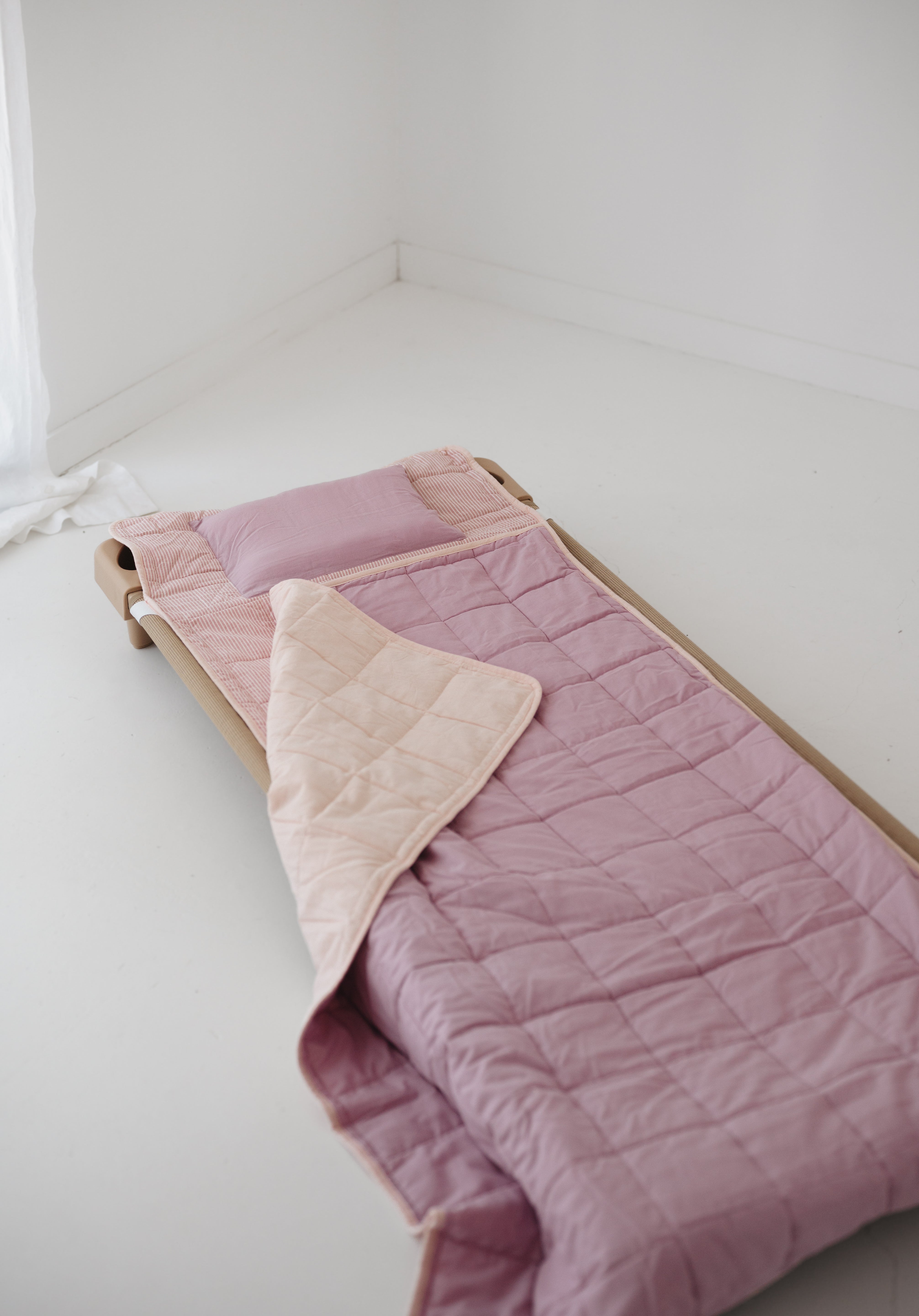 A soft purple and dusty pink stripe quilted daycare bedding nap mat with a soft peach underside on quilt. It has a built in matching pillowcase, pillow insert, name tag and carry handle. The daycare bedding nap mat is laid out on a daycare stacker bed frame against a white background.