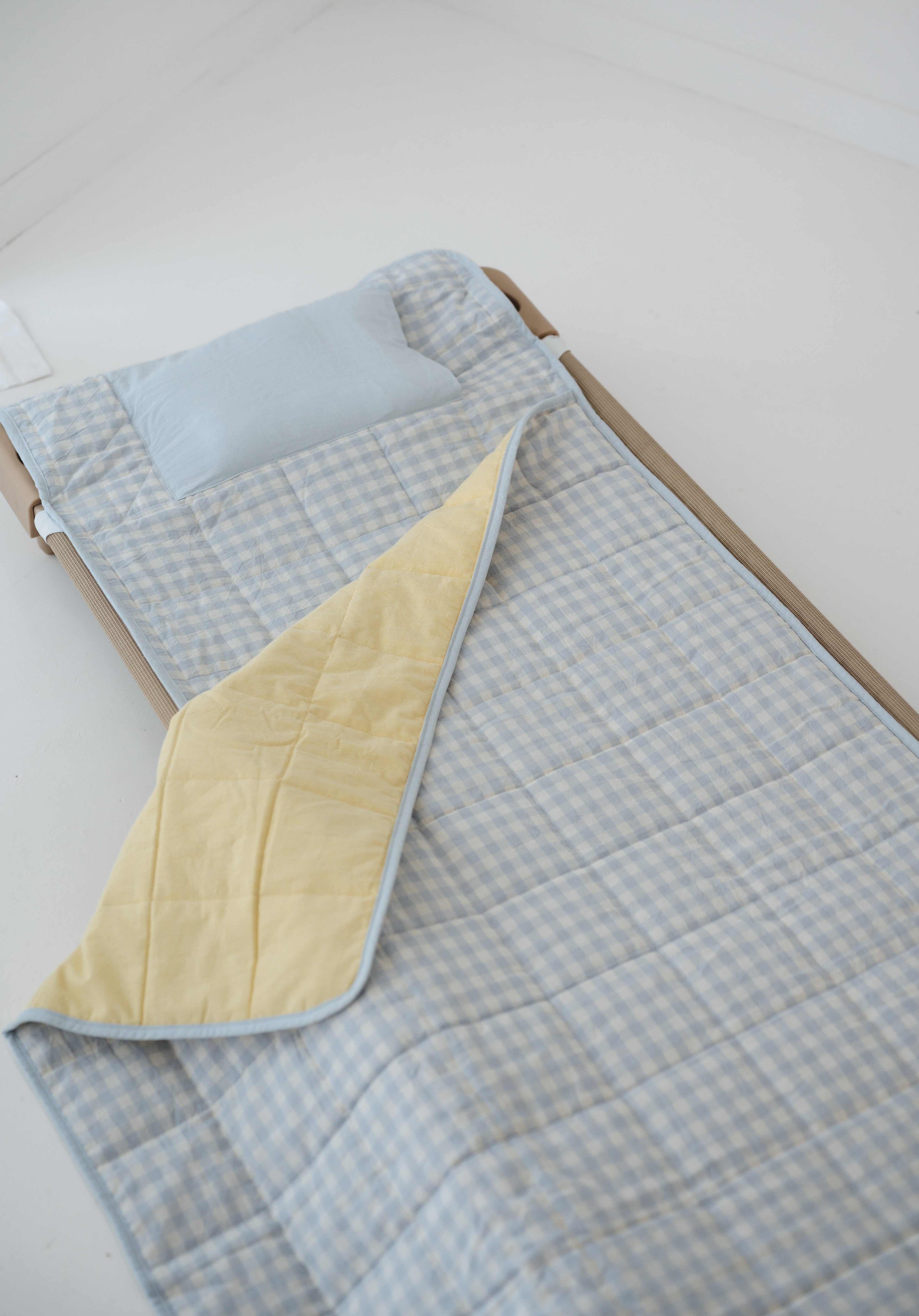 A light blue and white gingham quilted daycare bedding nap mat with soft yellow on the underside of the blanket. It has a built in matching pillowcase, pillow insert, name tag and carry handle. The daycare bedding nap mat is laid out on a daycare stacker bed frame against a white background.
