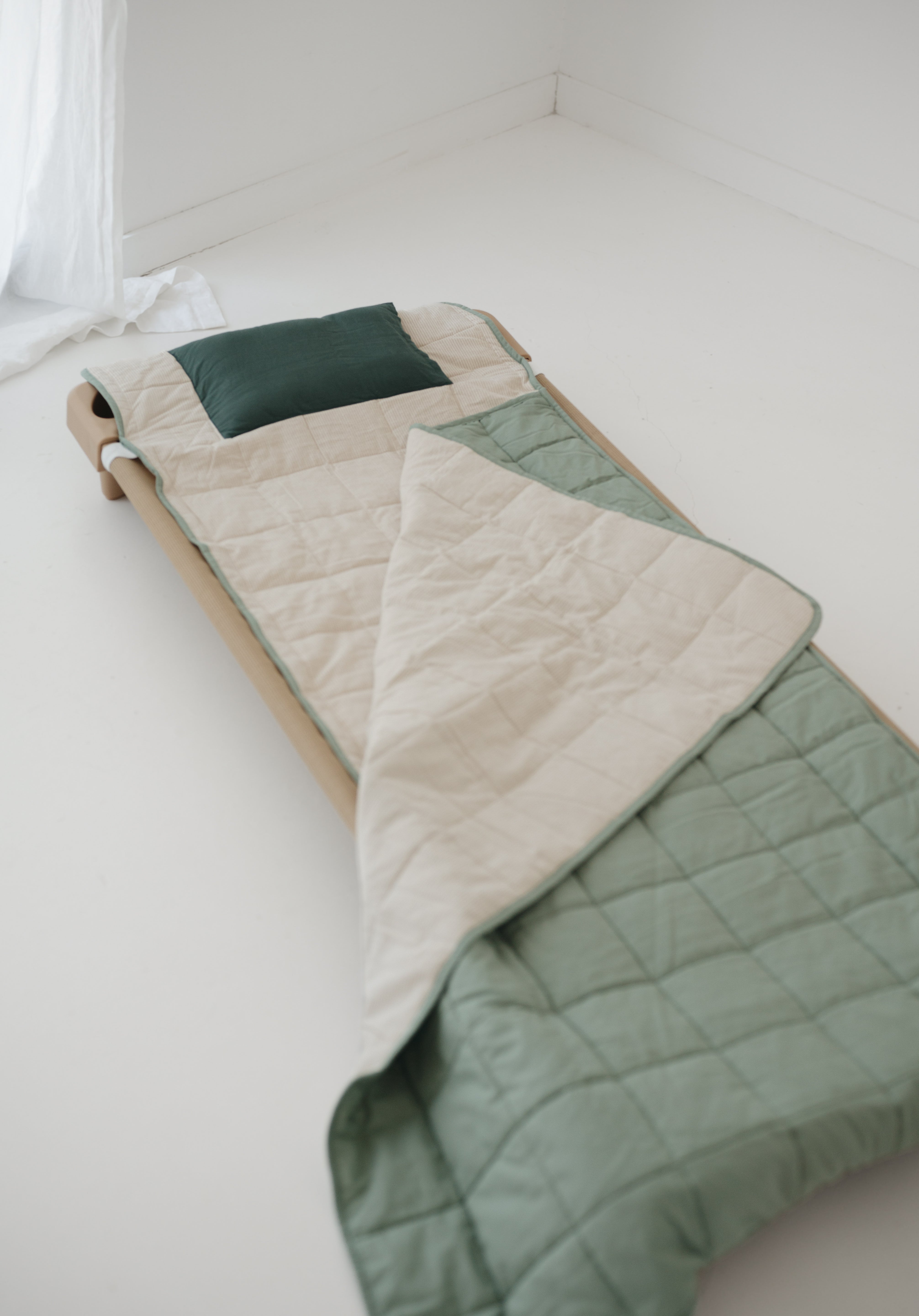 A olive green and beige stripe quilted daycare bedding nap mat with a dark green pillowcase attached. It has a built in matching pillowcase, pillow insert, name tag and carry handle. The daycare bedding nap mat is laid out on a daycare stacker bed frame against a white background.