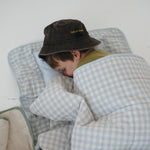 A light blue and white gingham quilted daycare bedding nap mat with soft yellow on the underside of the blanket. It has a built in matching pillowcase, pillow insert, name tag and carry handle. The daycare bedding nap mat is laid out on the floor being used a sleeping bag for a child and photographed with a white background.