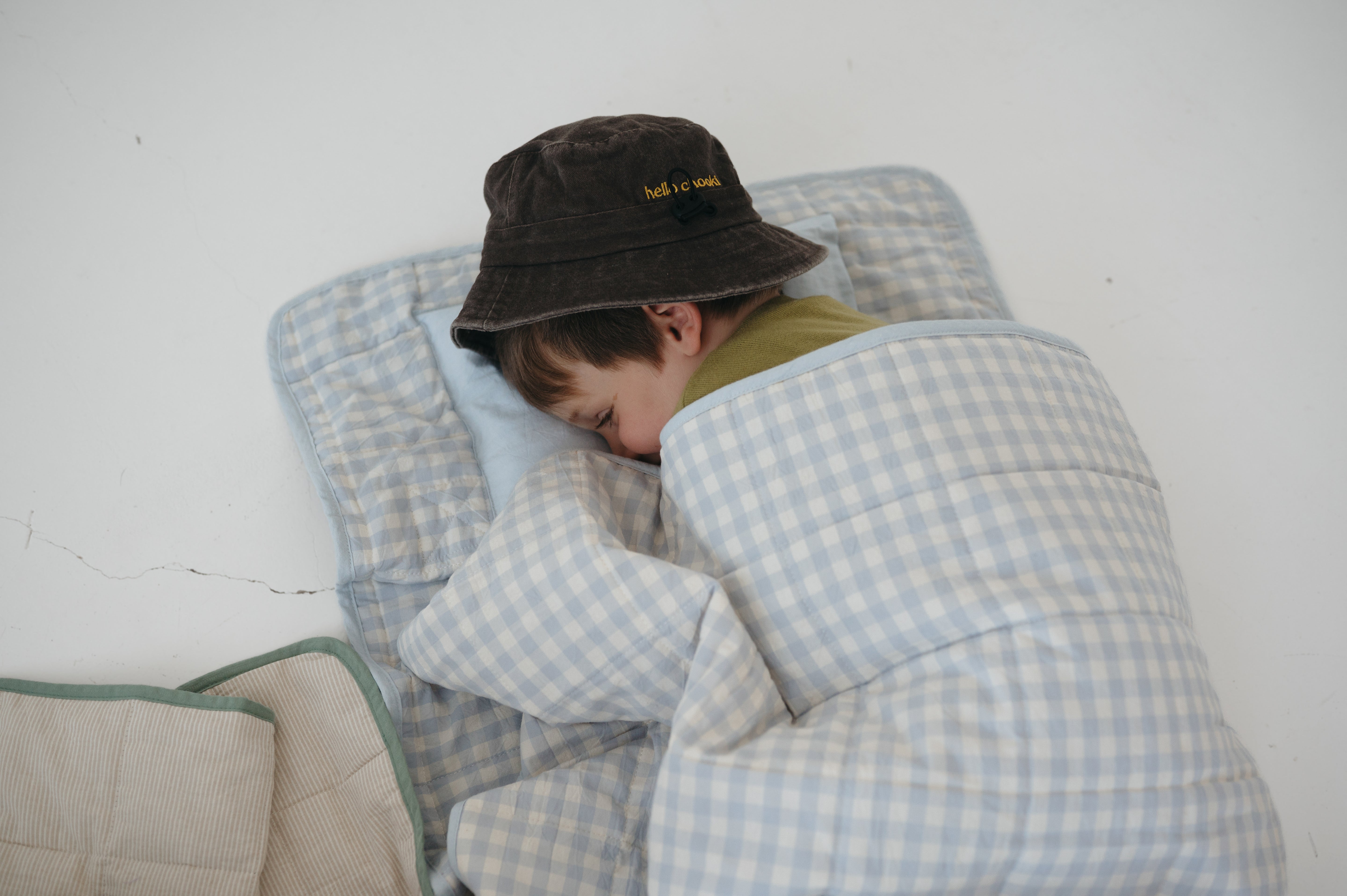 A light blue and white gingham quilted daycare bedding nap mat with soft yellow on the underside of the blanket. It has a built in matching pillowcase, pillow insert, name tag and carry handle. The daycare bedding nap mat is laid out on the floor being used a sleeping bag for a child and photographed with a white background.