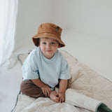 A olive green and beige stripe quilted daycare bedding nap mat with a dark green pillowcase attached. It has a built in matching pillowcase, pillow insert, name tag and carry handle. The daycare bedding nap mat is laid out on the floor being used as a sleeping bag with a toddler posing for the photo with a white background and wearing a Hello Chooki brown washed cotton daycare bucket hat.
