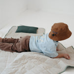 A olive green and beige stripe quilted daycare bedding nap mat with a dark green pillowcase attached. It has a built in matching pillowcase, pillow insert, name tag and carry handle. The daycare bedding nap mat is laid out on the floor with a toddler laying down and playing on the mat whilst wearing a Hello Chooki brown washed cotton daycare bucket hat, photographed with a white background.