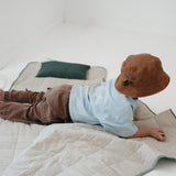 A olive green and beige stripe quilted daycare bedding nap mat with a dark green pillowcase attached. It has a built in matching pillowcase, pillow insert, name tag and carry handle. The daycare bedding nap mat is laid out on the floor with a toddler laying down and playing on the mat whilst wearing a Hello Chooki brown washed cotton daycare bucket hat, photographed with a white background.