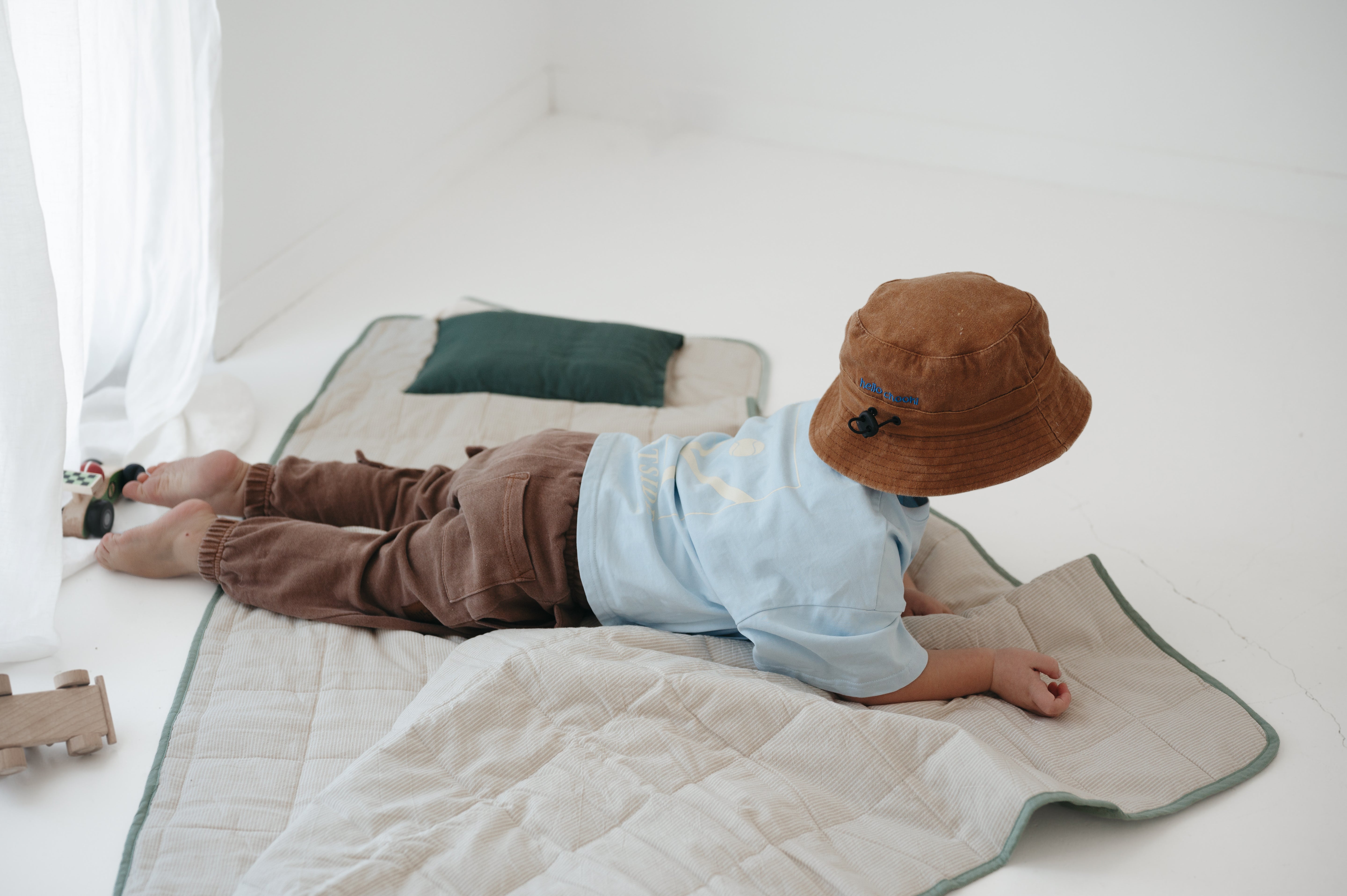 A olive green and beige stripe quilted daycare bedding nap mat with a dark green pillowcase attached. It has a built in matching pillowcase, pillow insert, name tag and carry handle. The daycare bedding nap mat is laid out on the floor with a toddler laying down and playing on the mat whilst wearing a Hello Chooki brown washed cotton daycare bucket hat, photographed with a white background.