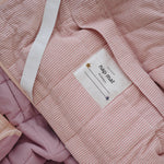 A soft purple and dusty pink stripe quilted daycare bedding nap mat with a soft peach underside on quilt. It has a built in matching pillowcase, pillow insert, name tag and carry handle. The daycare bedding nap mat is laid out showing the name tag, carry handle and elastic straps that help secure it to a daycare stacker bed and rolled up when not in use.