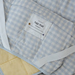 A light blue and white gingham quilted daycare bedding nap mat with soft yellow on the underside of the blanket. It has a built in matching pillowcase, pillow insert, name tag and carry handle. The daycare bedding nap mat is laid out showing in detail the name tag, carry handle and elastic straps that help secure it to the daycare stacker bed and also keeping it rolled up when not in use.