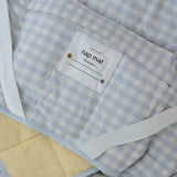 A light blue and white gingham quilted daycare bedding nap mat with soft yellow on the underside of the blanket. It has a built in matching pillowcase, pillow insert, name tag and carry handle. The daycare bedding nap mat is laid out showing in detail the name tag, carry handle and elastic straps that help secure it to the daycare stacker bed and also keeping it rolled up when not in use.