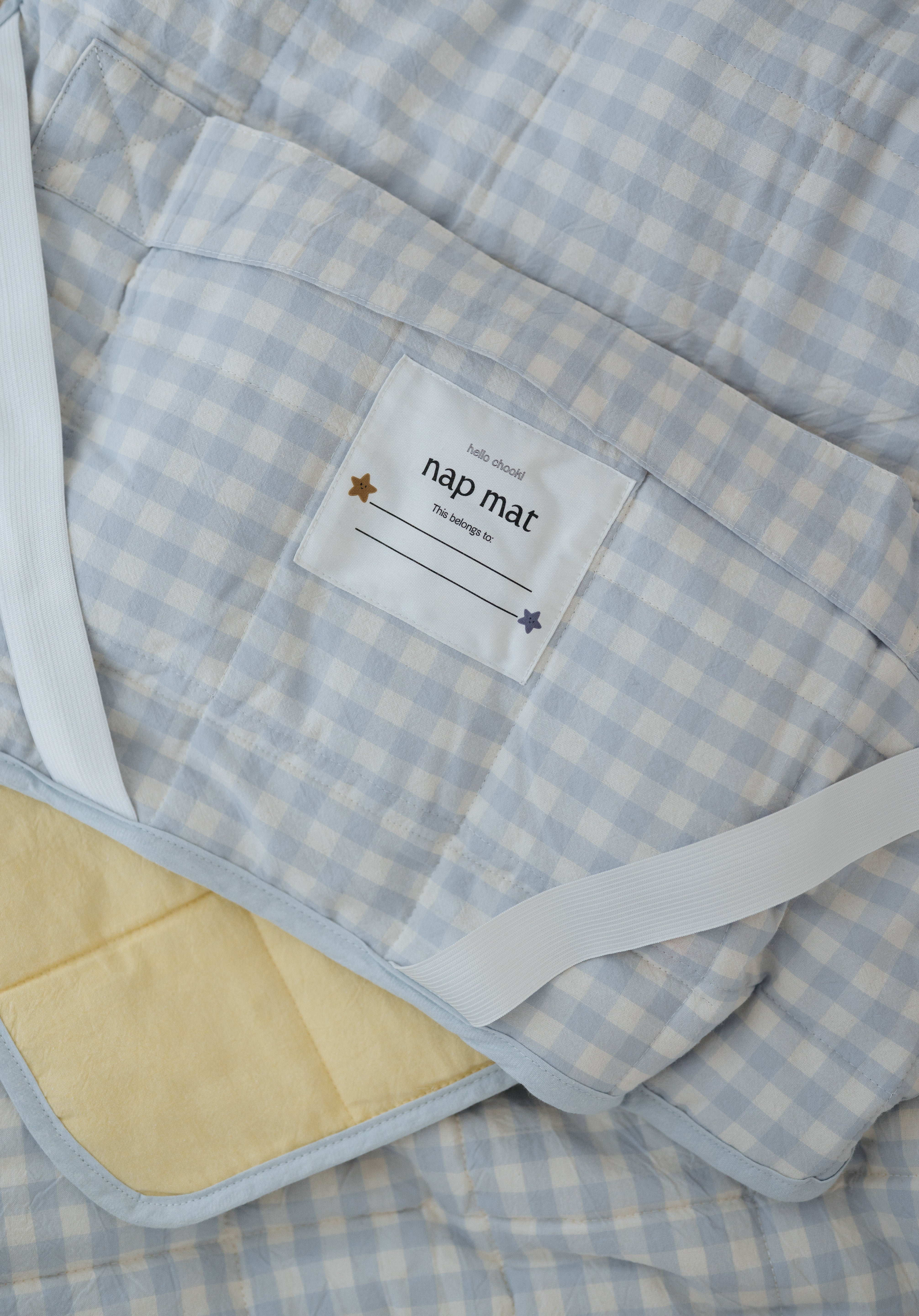 A light blue and white gingham quilted daycare bedding nap mat with soft yellow on the underside of the blanket. It has a built in matching pillowcase, pillow insert, name tag and carry handle. The daycare bedding nap mat is laid out showing in detail the name tag, carry handle and elastic straps that help secure it to the daycare stacker bed and also keeping it rolled up when not in use.
