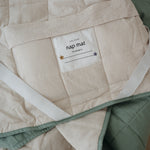 A olive green and beige stripe quilted daycare bedding nap mat with a dark green pillowcase attached. It has a built in matching pillowcase, pillow insert, name tag and carry handle. The daycare bedding nap mat is laid out showing a close up of the attached name tag, carry handle and elastic straps that help secure it to a daycare stacker bed and keeping it rolled up when not in use.