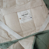 A olive green and beige stripe quilted daycare bedding nap mat with a dark green pillowcase attached. It has a built in matching pillowcase, pillow insert, name tag and carry handle. The daycare bedding nap mat is laid out showing a close up of the attached name tag, carry handle and elastic straps that help secure it to a daycare stacker bed and keeping it rolled up when not in use.