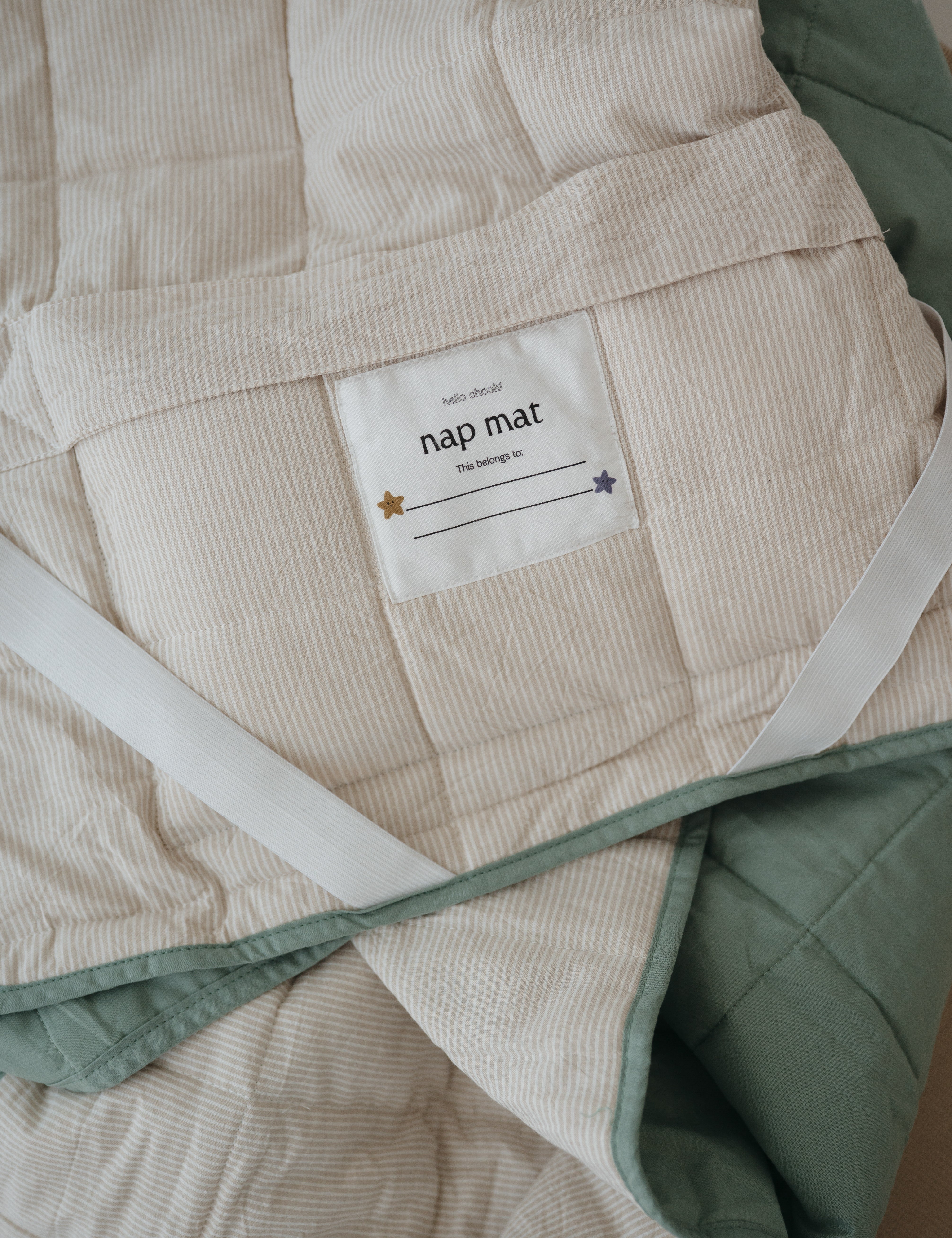 A olive green and beige stripe quilted daycare bedding nap mat with a dark green pillowcase attached. It has a built in matching pillowcase, pillow insert, name tag and carry handle. The daycare bedding nap mat is laid out showing a close up of the attached name tag, carry handle and elastic straps that help secure it to a daycare stacker bed and keeping it rolled up when not in use.