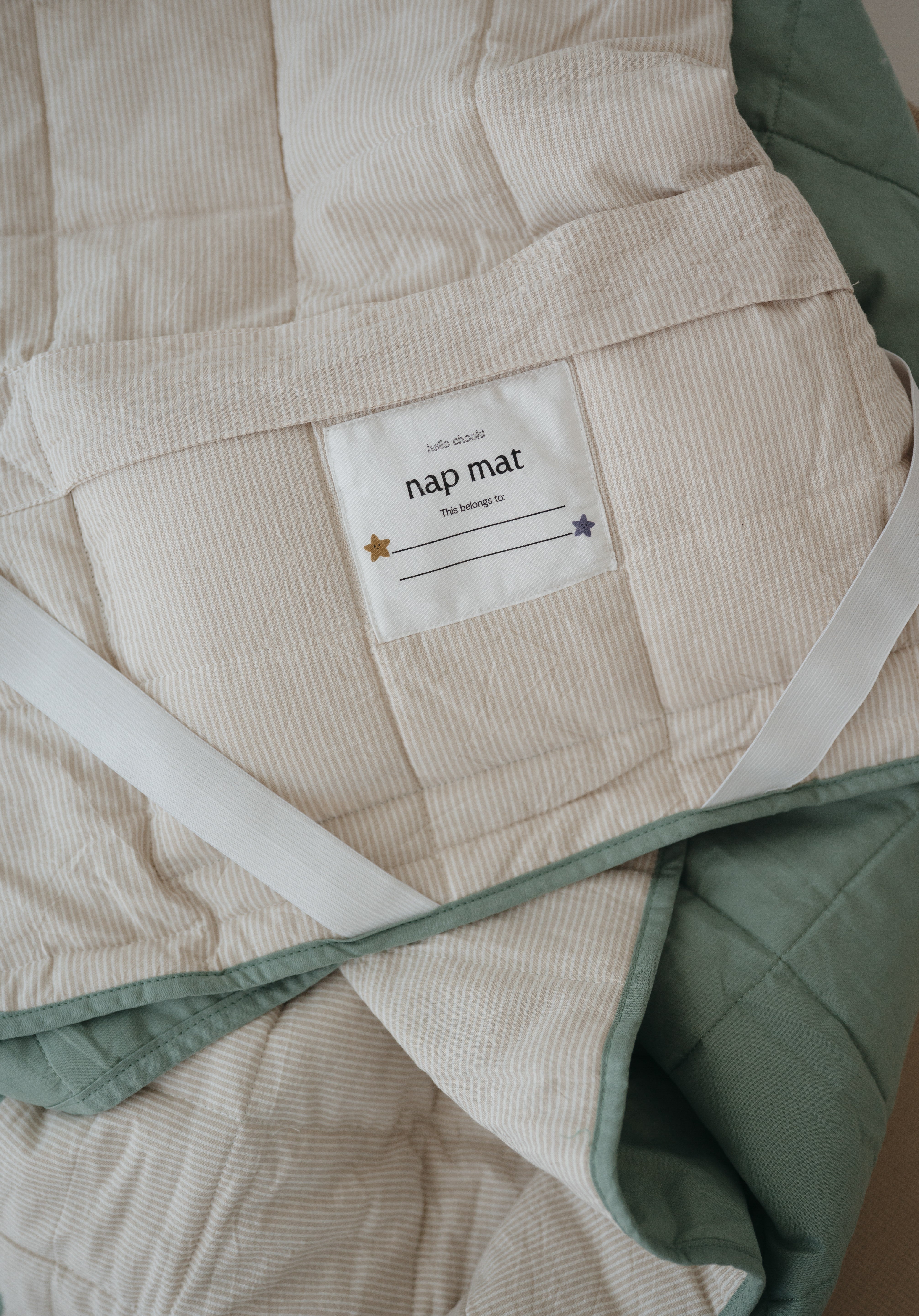 A olive green and beige stripe quilted daycare bedding nap mat with a dark green pillowcase attached. It has a built in matching pillowcase, pillow insert, name tag and carry handle. The daycare bedding nap mat is laid out showing a close up of the attached name tag, carry handle and elastic straps that help secure it to a daycare stacker bed and keeping it rolled up when not in use.