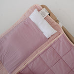 A soft purple and dusty pink stripe quilted daycare bedding nap mat with a soft peach underside on quilt. It has a built in matching pillowcase, pillow insert, name tag and carry handle. The daycare bedding nap mat is laid out on a daycare stacker bed frame against a white background showing the removable pillow insert.