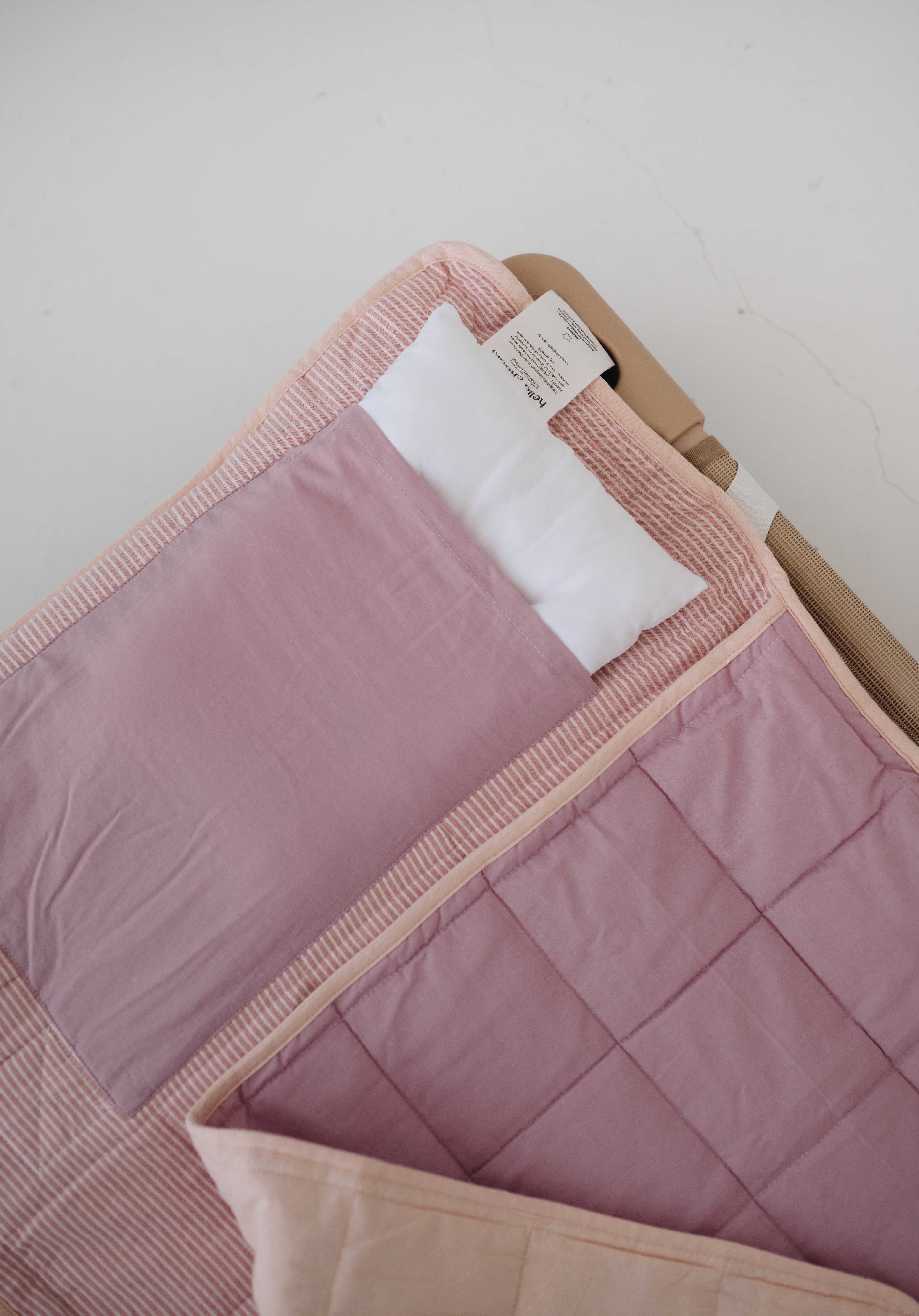 A soft purple and dusty pink stripe quilted daycare bedding nap mat with a soft peach underside on quilt. It has a built in matching pillowcase, pillow insert, name tag and carry handle. The daycare bedding nap mat is laid out on a daycare stacker bed frame against a white background showing the removable pillow insert.