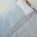 A light blue and white gingham quilted daycare bedding nap mat with soft yellow on the underside of the blanket. It has a built in matching pillowcase, pillow insert, name tag and carry handle. The daycare bedding nap mat is laid out on a daycare stacker bed frame against a white background showing that the daycare nap has a pillow insert included.