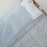 A light blue and white gingham quilted daycare bedding nap mat with soft yellow on the underside of the blanket. It has a built in matching pillowcase, pillow insert, name tag and carry handle. The daycare bedding nap mat is laid out on a daycare stacker bed frame against a white background showing that the daycare nap has a pillow insert included.