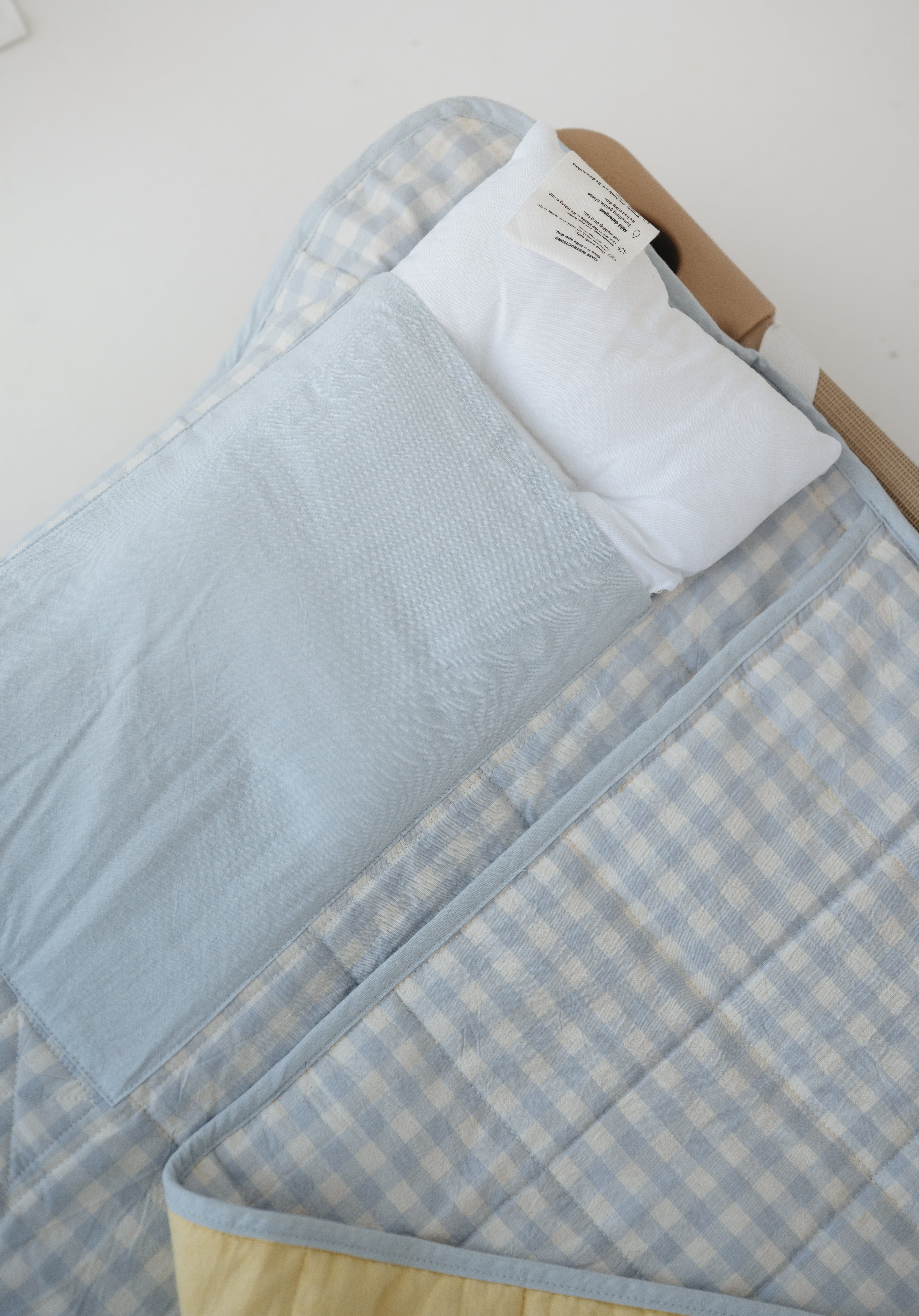 A light blue and white gingham quilted daycare bedding nap mat with soft yellow on the underside of the blanket. It has a built in matching pillowcase, pillow insert, name tag and carry handle. The daycare bedding nap mat is laid out on a daycare stacker bed frame against a white background showing that the daycare nap has a pillow insert included.