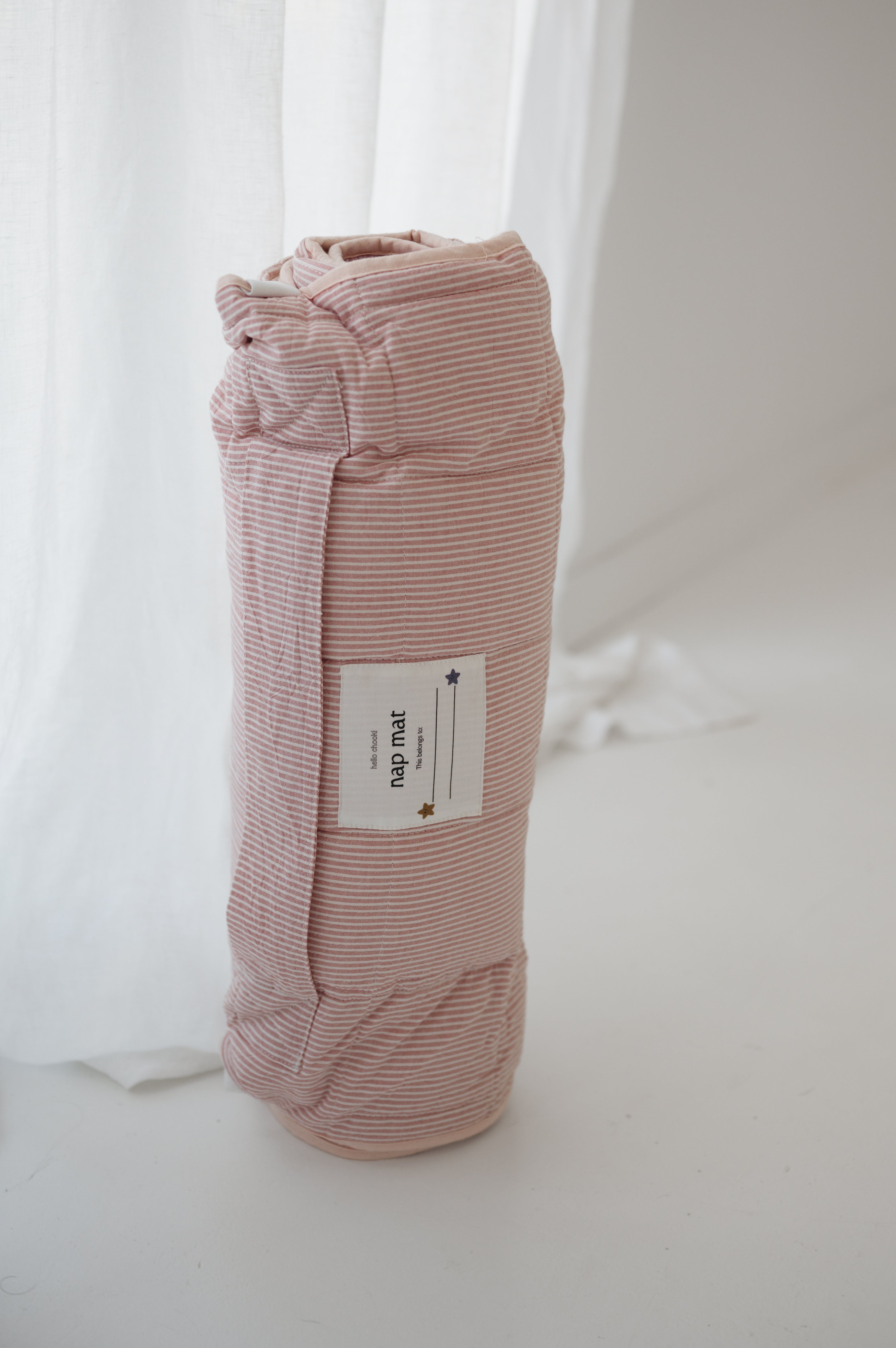A soft purple and dusty pink stripe quilted daycare bedding nap mat with a soft peach underside on quilt. It has a built in matching pillowcase, pillow insert, name tag and carry handle. The daycare bedding nap mat is rolled up against a white background.
