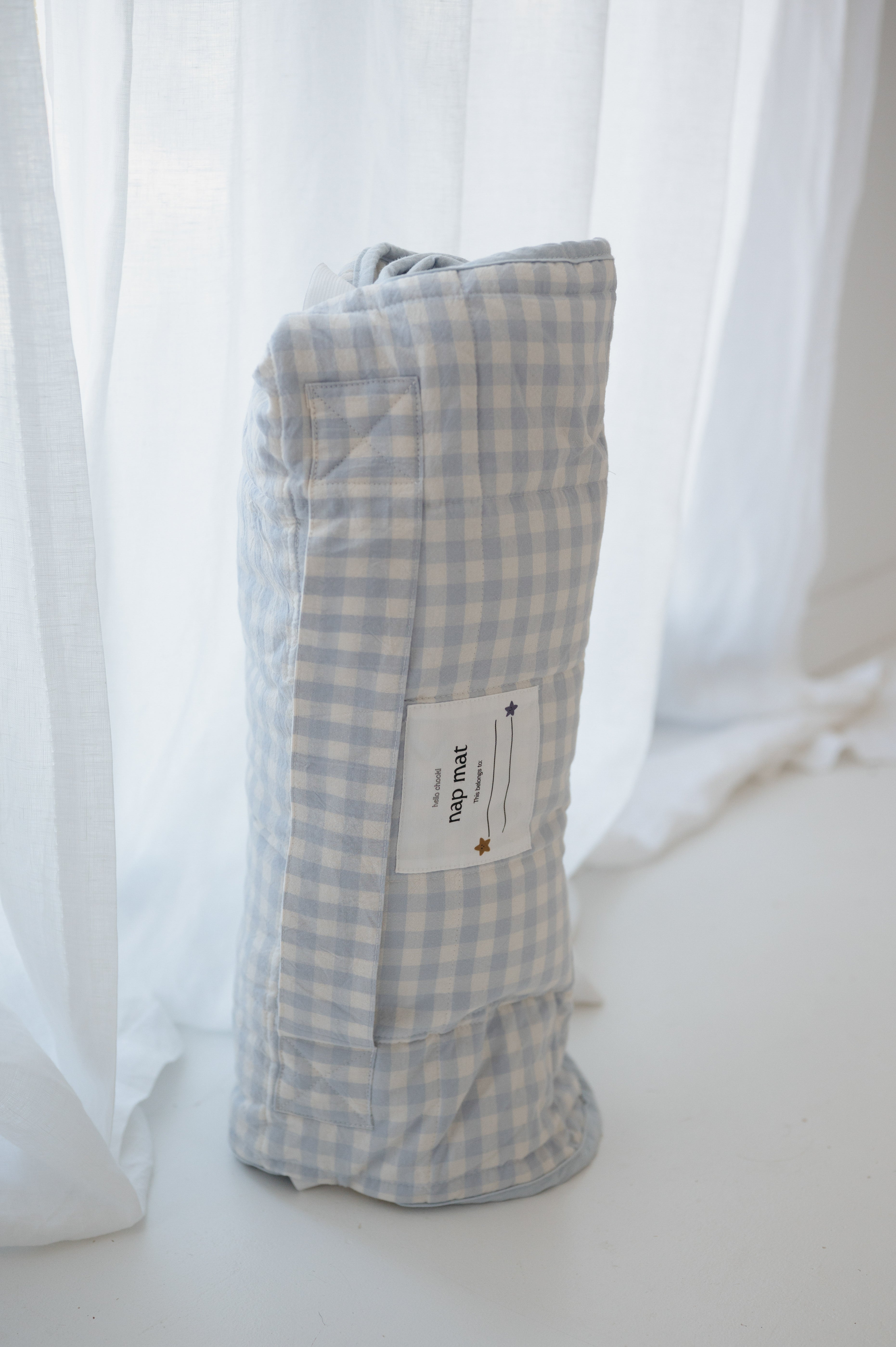 A light blue and white gingham quilted daycare bedding nap mat with soft yellow on the underside of the blanket. It has a built in matching pillowcase, pillow insert, name tag and carry handle. The daycare bedding nap mat is rolled up showing how it is packed away not in use and photographed against a white background.