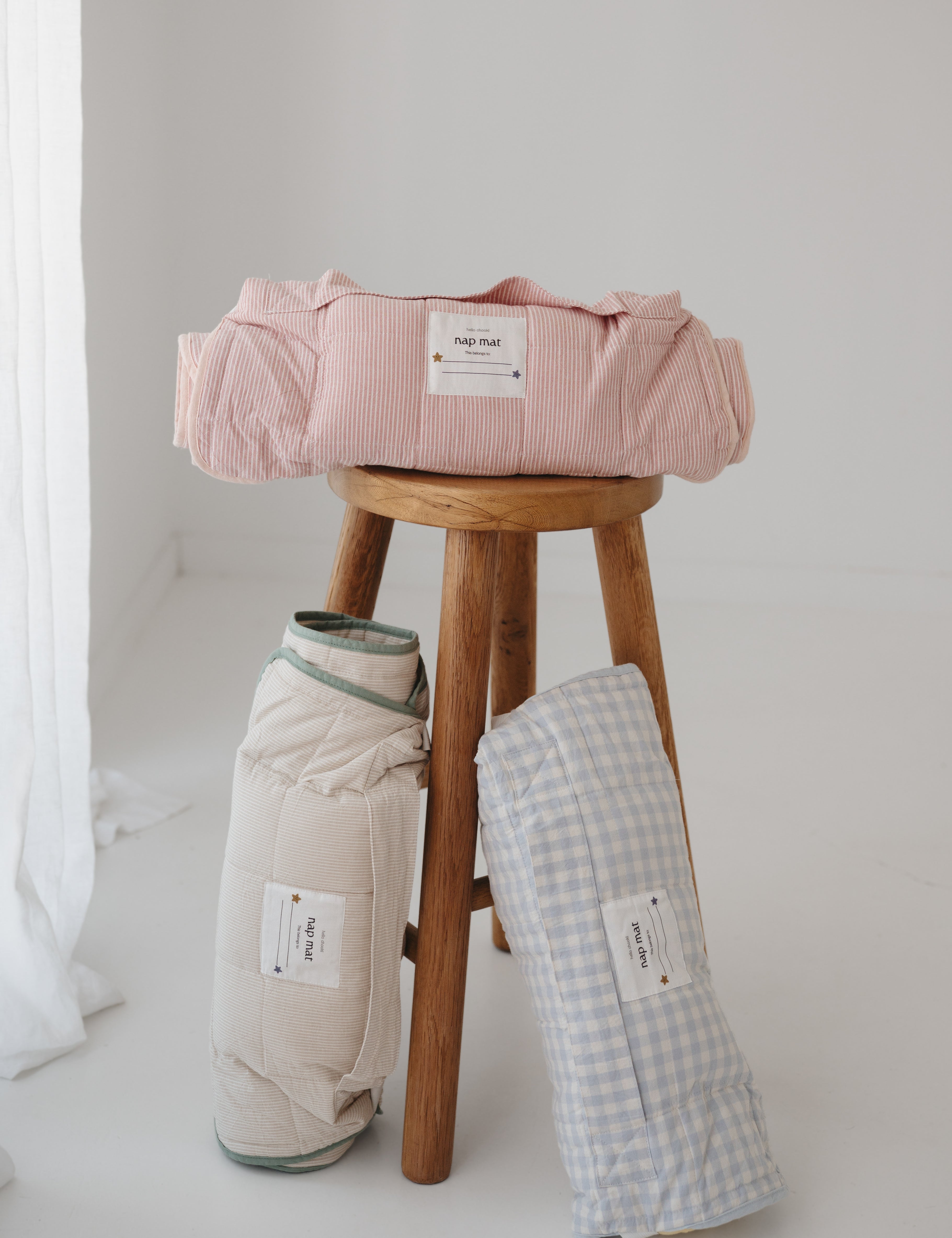 A olive green and beige stripe quilted daycare bedding nap mat with a dark green pillowcase attached. It has a built in matching pillowcase, pillow insert, name tag and carry handle. The daycare bedding nap mat is rolled up against a timber stool next to alternative prints from the daycare bedding nap mat collection, photographed with a white background.
