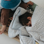 A light blue and white gingham quilted daycare bedding nap mat with soft yellow on the underside of the blanket. It has a built in matching pillowcase, pillow insert, name tag and carry handle. The daycare bedding nap mat is laid out on a daycare stacker bed frame with children using it to play and set against a white background.