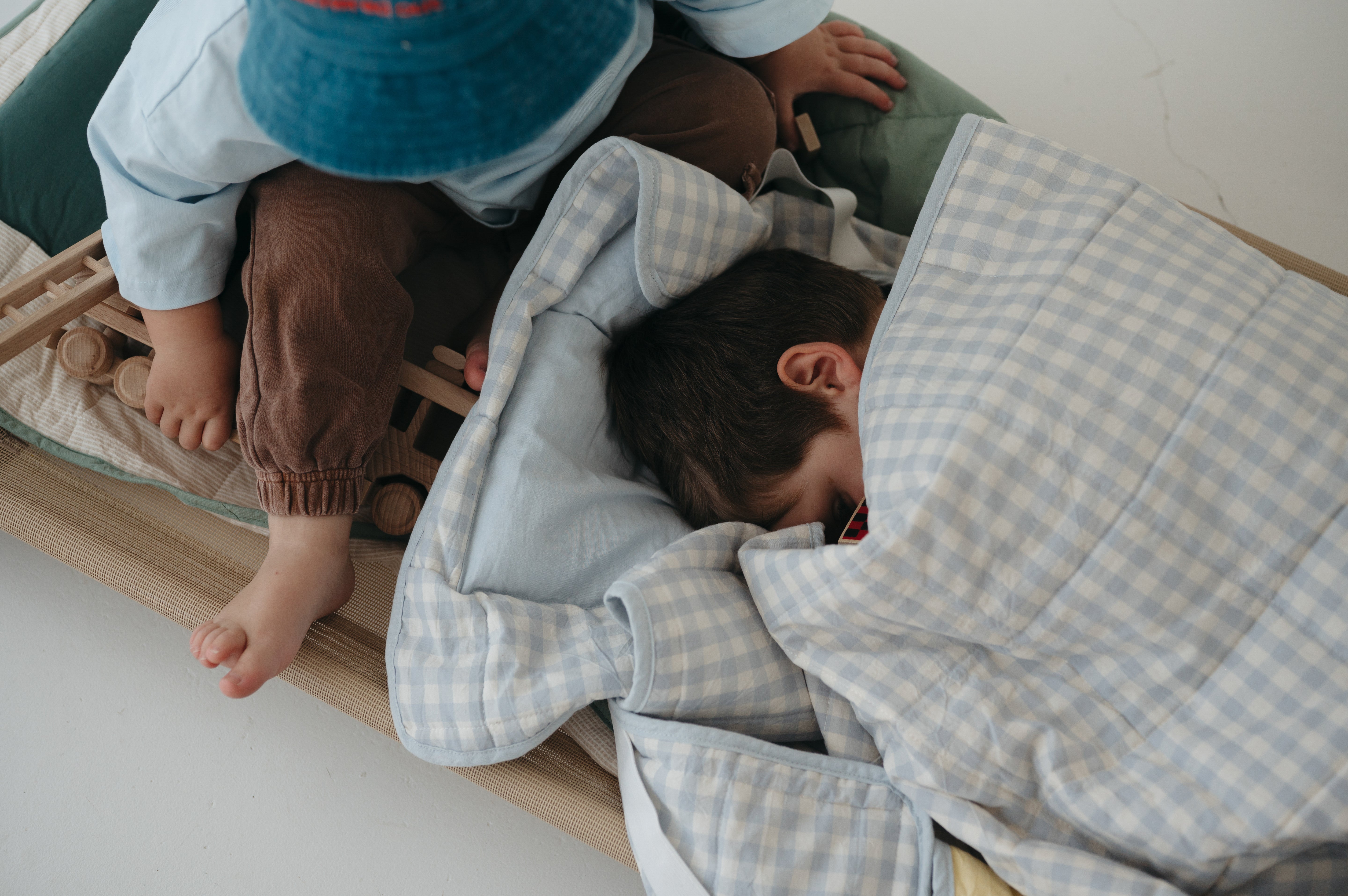 A light blue and white gingham quilted daycare bedding nap mat with soft yellow on the underside of the blanket. It has a built in matching pillowcase, pillow insert, name tag and carry handle. The daycare bedding nap mat is laid out on a daycare stacker bed frame with children using it to play and set against a white background.