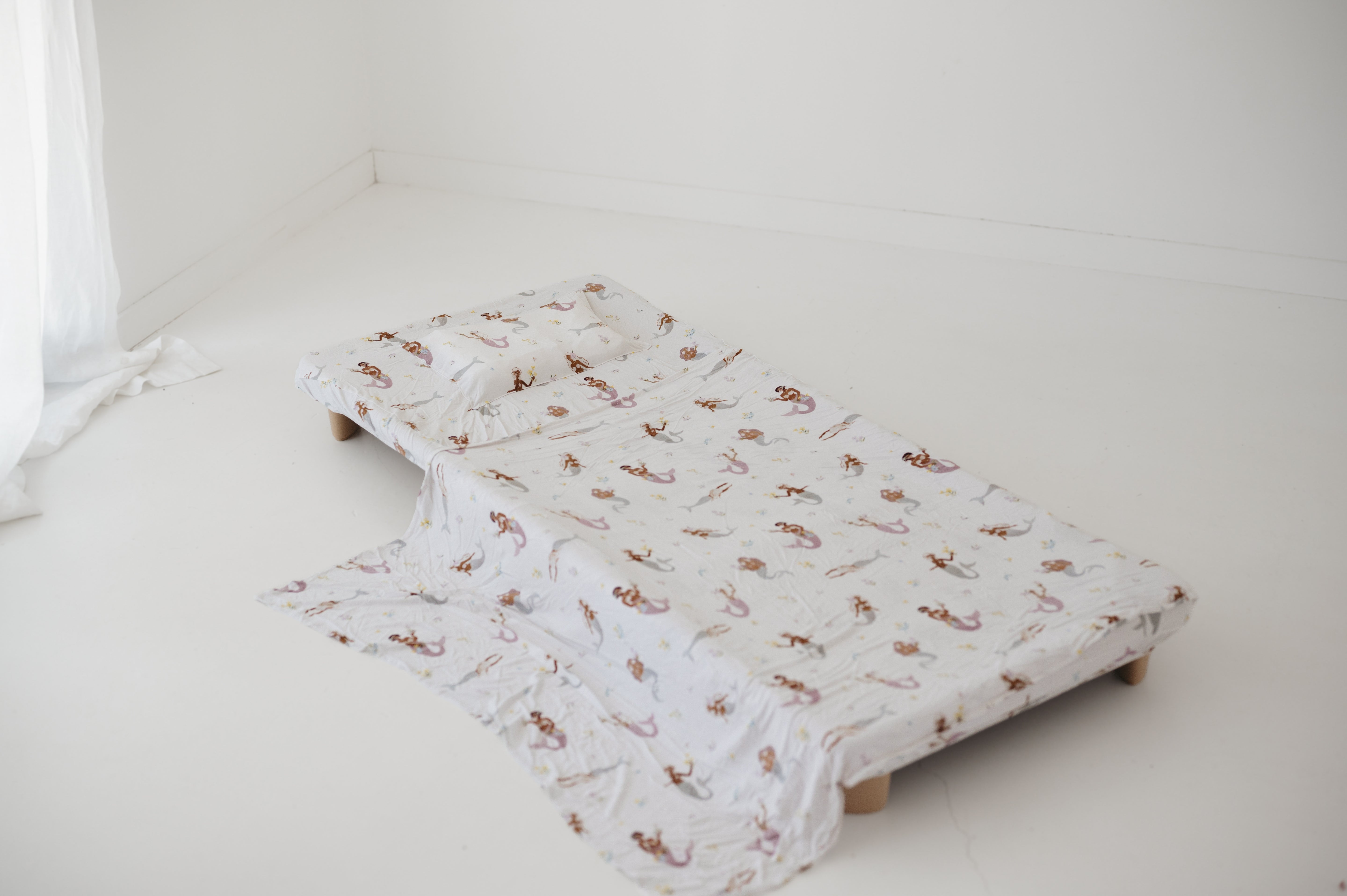 A white and multicolour bohemian style mermaid sheet set laid out onto a daycare stacker bed. The print features hand painted mermaids of different tones, with various colour tails and hair colours including soft blue, soft purple and soft pink. The mermaids have flowers in their hair and hands with same flowers scattered throughout. The products are photographed with a white background.
