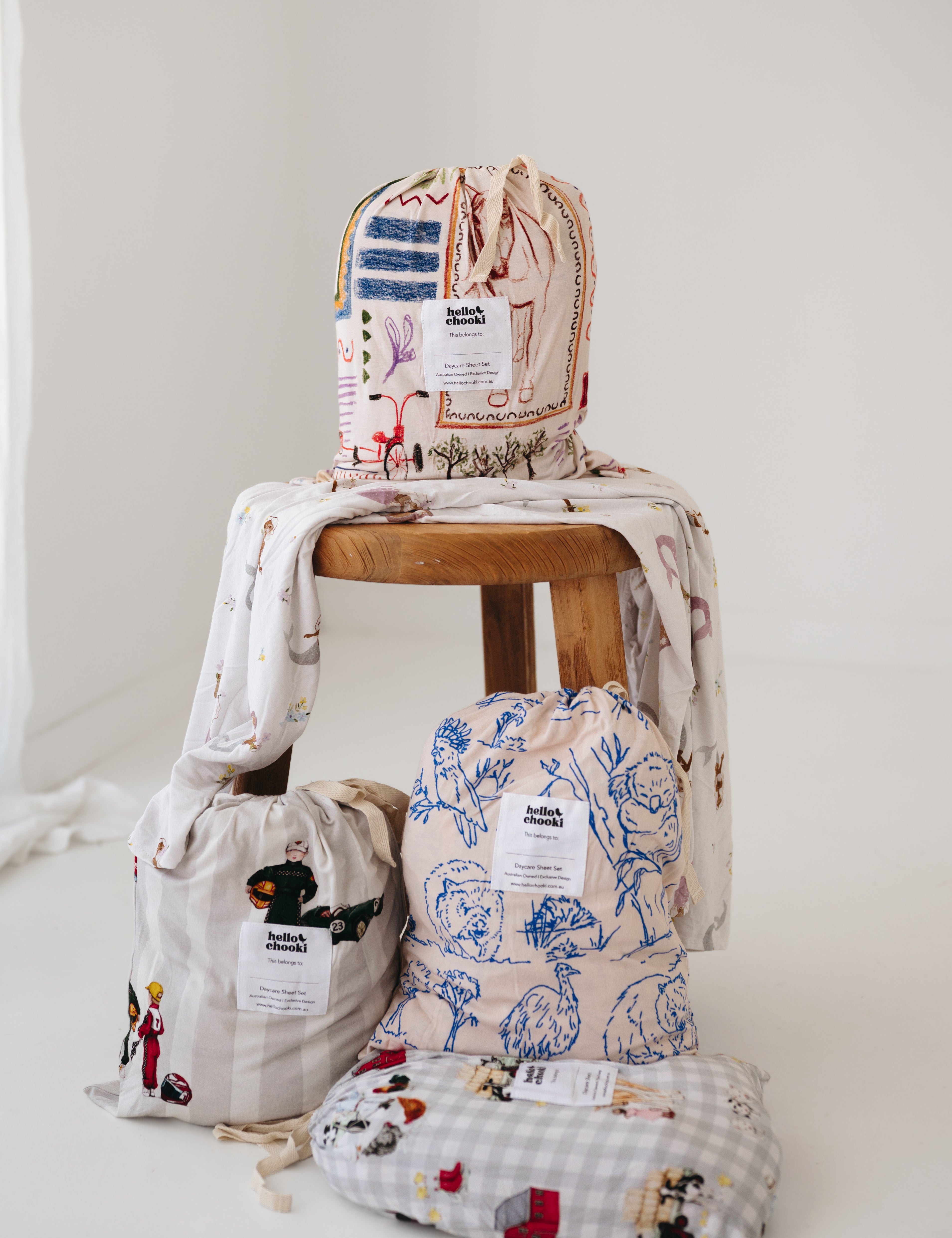 A soft beige and multicolour sheet set packaged in its matching drawstring storage bag sitting on a round wooden stool next to alternative daycare sheet sets from the Hello Chooki collection. The print features hand drawn geometric. The products are photographed with a white background. shapes with nostalgic images including a horse, vintage pedal bike, tent and camp fire, person fishing along with native trees, flowers and animals. Photographed on a white background.