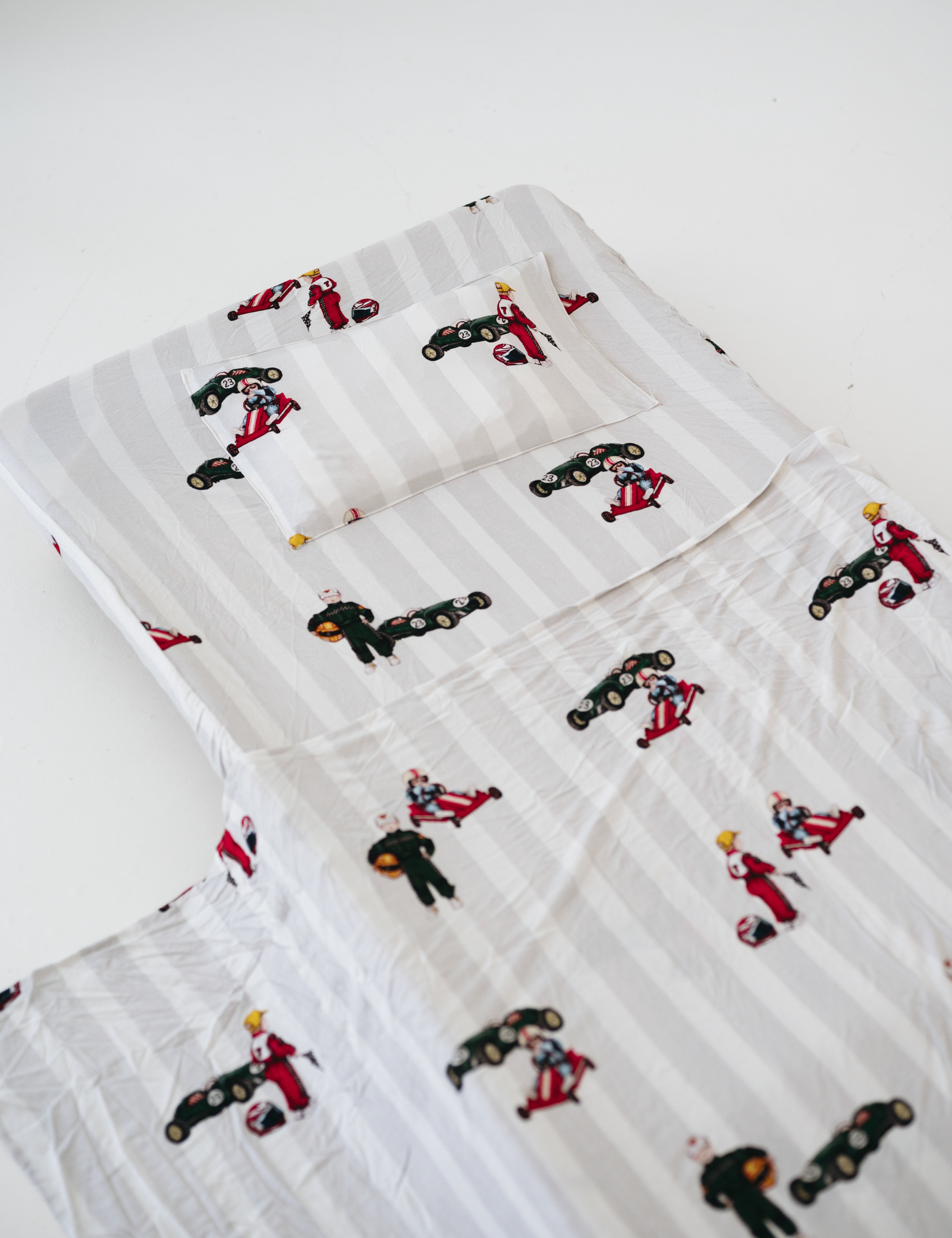 A white and soft grey striped daycare sheet set laid out onto a daycare stacker bed. The print features vintage hand drawn boys dressed in vintage race overalls and helmet next to a vintage race car and driving a vintage go kart. The products are photographed with a white background.
