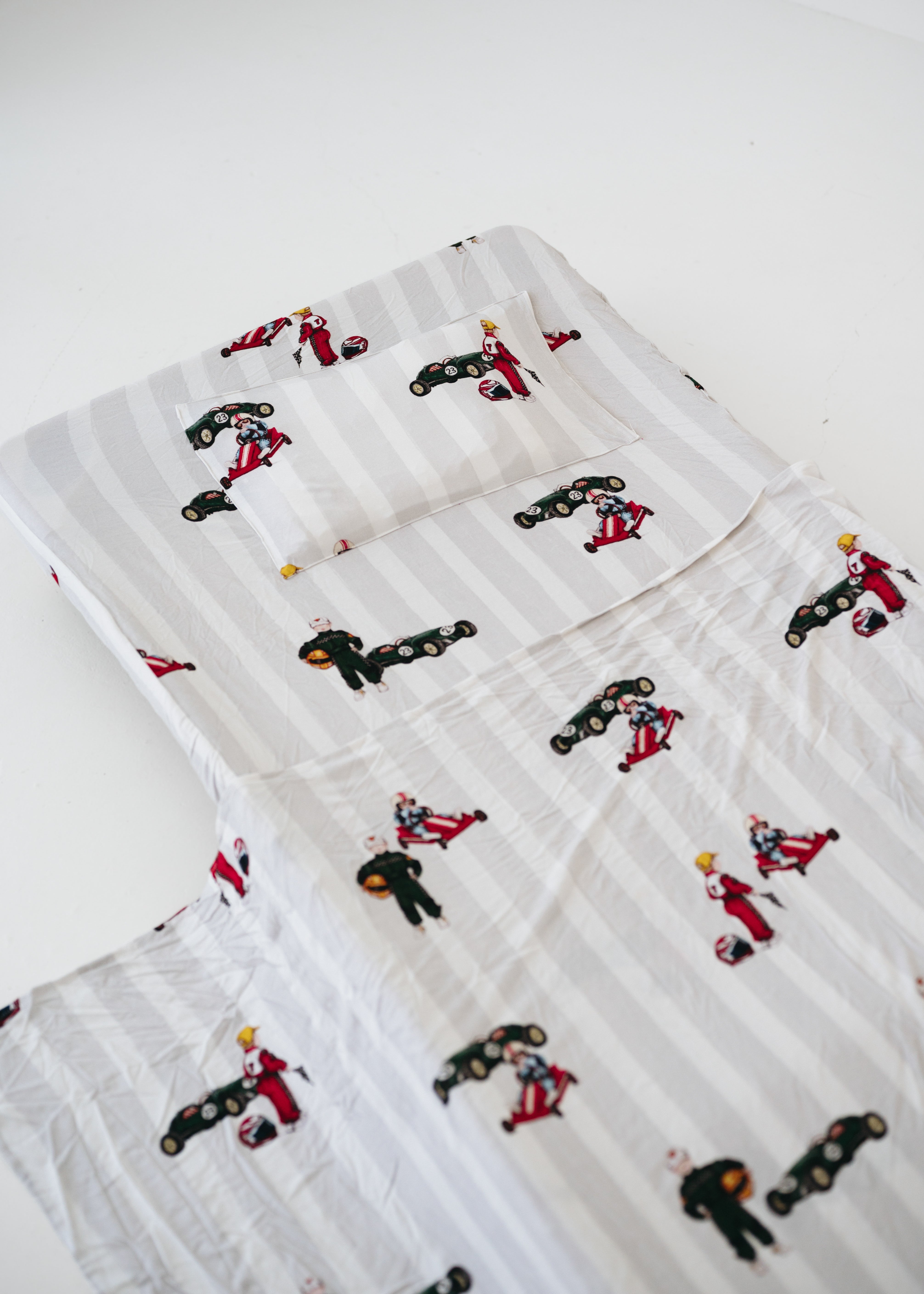 A white and soft grey striped daycare sheet set laid out onto a daycare stacker bed. The print features vintage hand drawn boys dressed in vintage race overalls and helmet next to a vintage race car and driving a vintage go kart. The products are photographed with a white background.