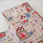 A soft beige and multicolour sheet set laid out onto a daycare stacker bed. The print features hand drawn geometric. The products are photographed with a white background. shapes with nostalgic images including a horse, vintage pedal bike, tent and camp fire, person fishing along with native trees, flowers and animals. Photographed on a white background.