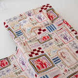 A soft beige and multicolour sheet set laid out onto a daycare stacker bed. The print features hand drawn geometric. The products are photographed with a white background. shapes with nostalgic images including a horse, vintage pedal bike, tent and camp fire, person fishing along with native trees, flowers and animals. Photographed on a white background.