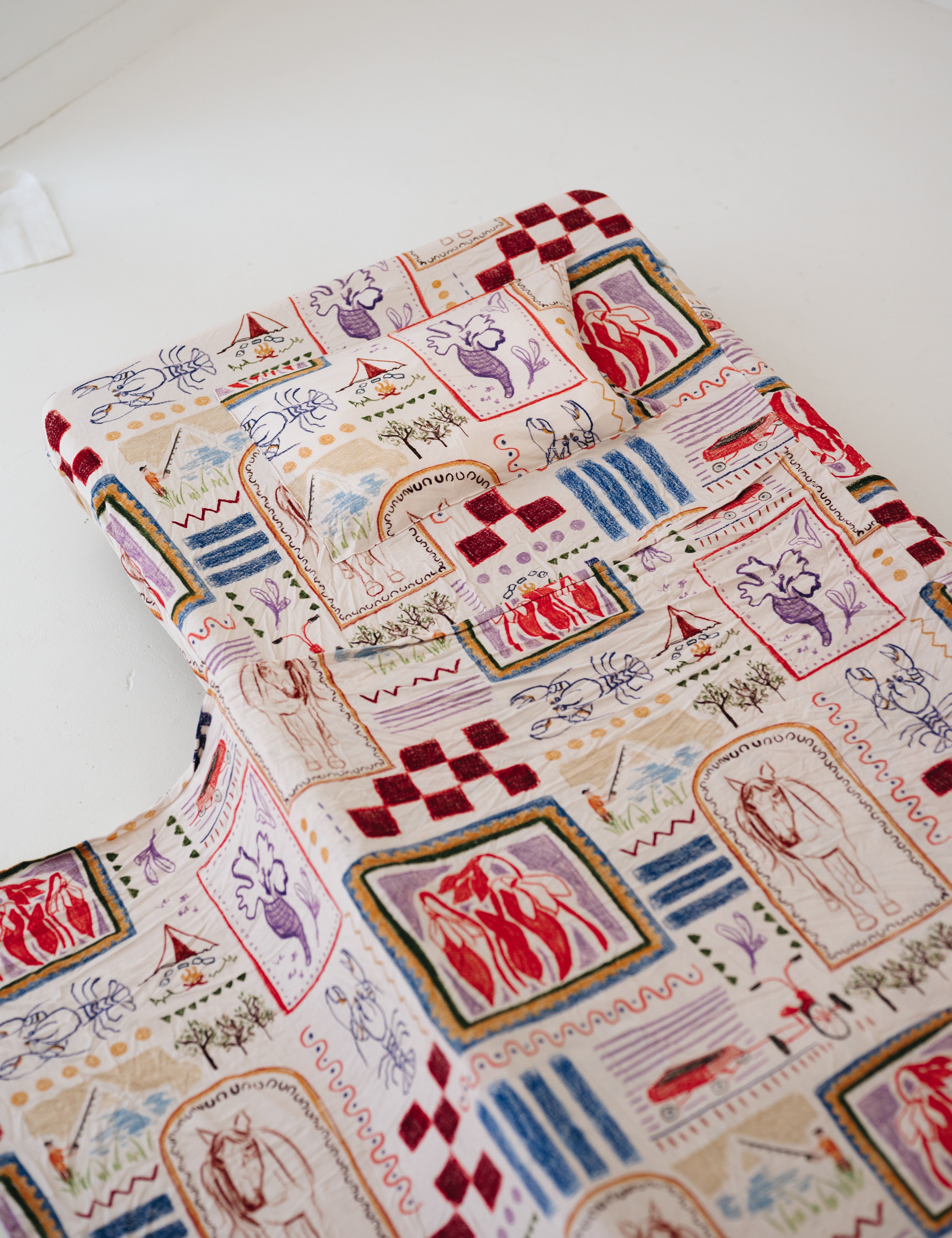 A soft beige and multicolour sheet set laid out onto a daycare stacker bed. The print features hand drawn geometric. The products are photographed with a white background. shapes with nostalgic images including a horse, vintage pedal bike, tent and camp fire, person fishing along with native trees, flowers and animals. Photographed on a white background.